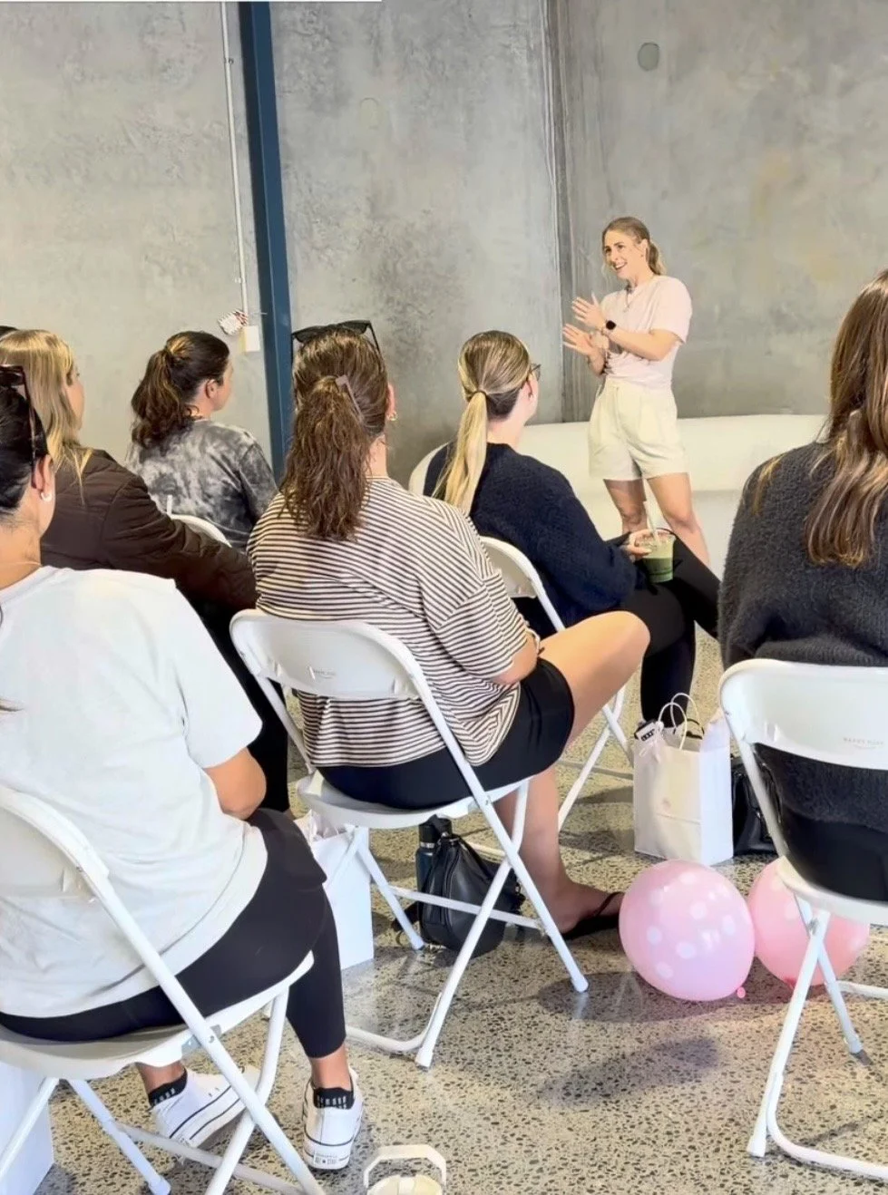 A woman (Kat Rogers - pelvic health physiotherapist) standing next to a group of seated women, giving a presentation or speech in a modern room with a concrete wall. Two pink balloons are on the floor at the front.