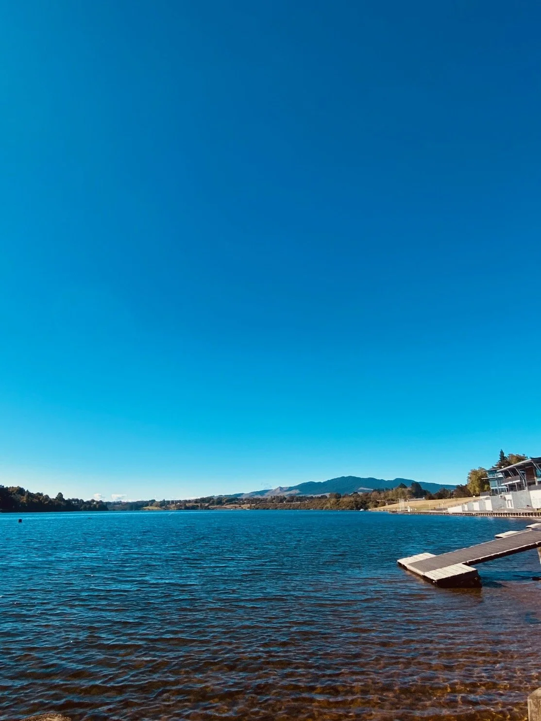 A calm Lake Karapiro in the Waikato, with a clear blue sky above, distant mountains on the horizon, and modern dock and house on the right side.