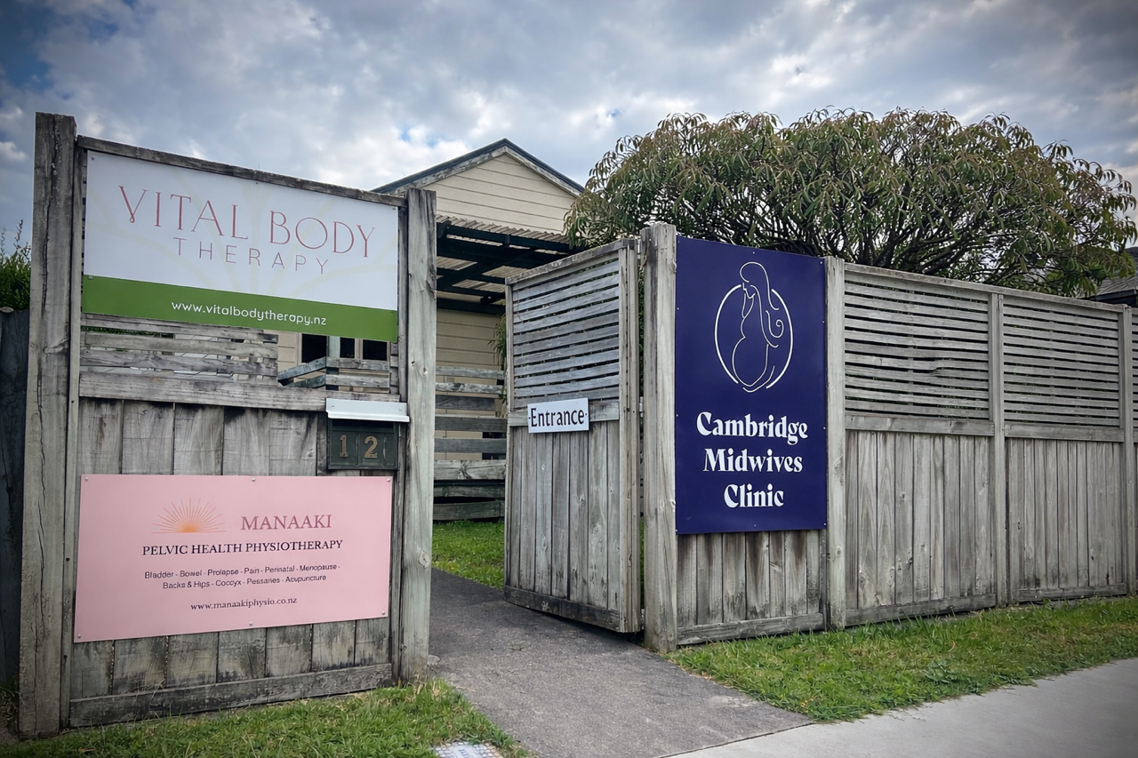 Entrance to Cambridge Midwives Clinic with wooden fence, signs for Vital Body Therapy and Manaaki Pelvic Health Physiotherapy, and a cloudy sky.