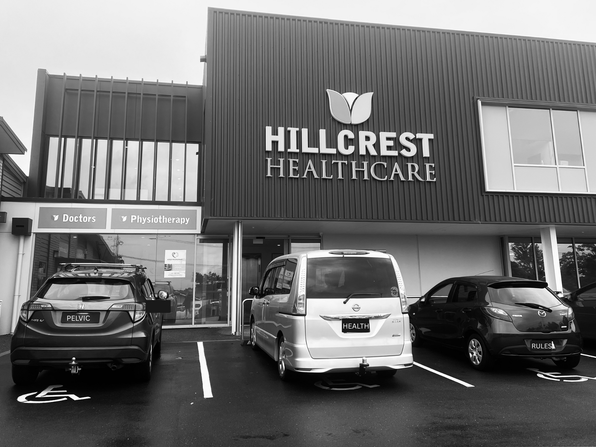 The exterior of Hillcrest Healthcare building with parking lot in front, featuring cars labeled 'Pelvic,' 'Health,' and 'Rules,' in black and white.