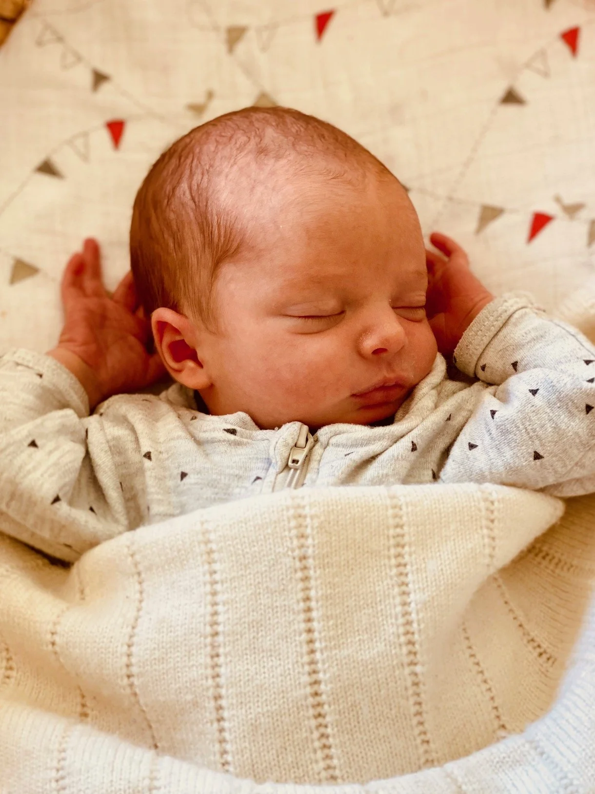 A sleeping newborn baby with reddish hair, dressed in a light-colored long sleeve shirt, lying on a blanket with red and gray triangle patterns.