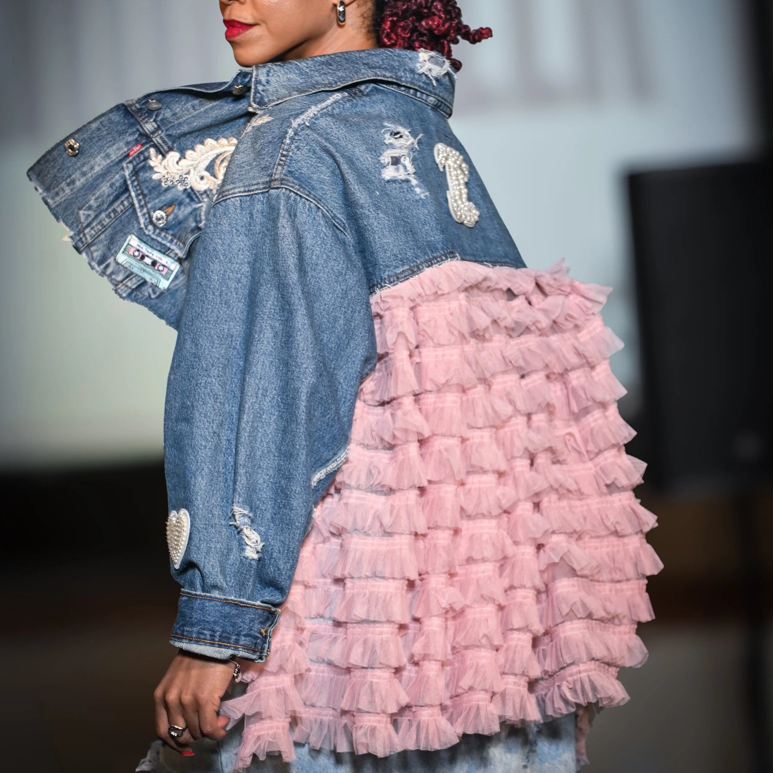 Person wearing a blue denim jacket with embroidered patches and rhinestones, layered with a pink ruffled fabric overlay on the back.