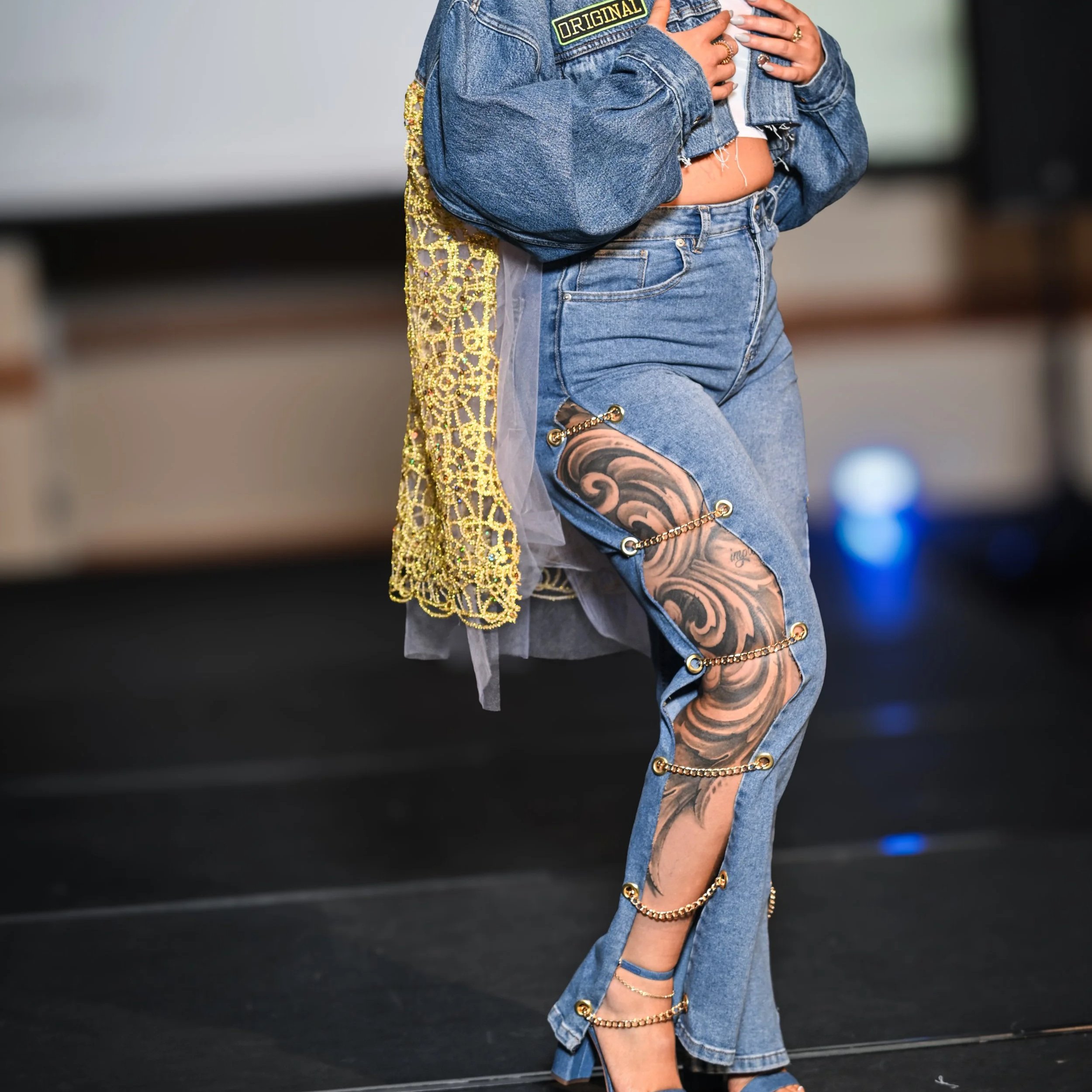 Close-up of a person wearing light blue ripped jeans decorated with gold chains, high-heeled shoes, a denim jacket with a patch that says 'Original', and a metallic gold lace and denim jacket, standing on a black surface.