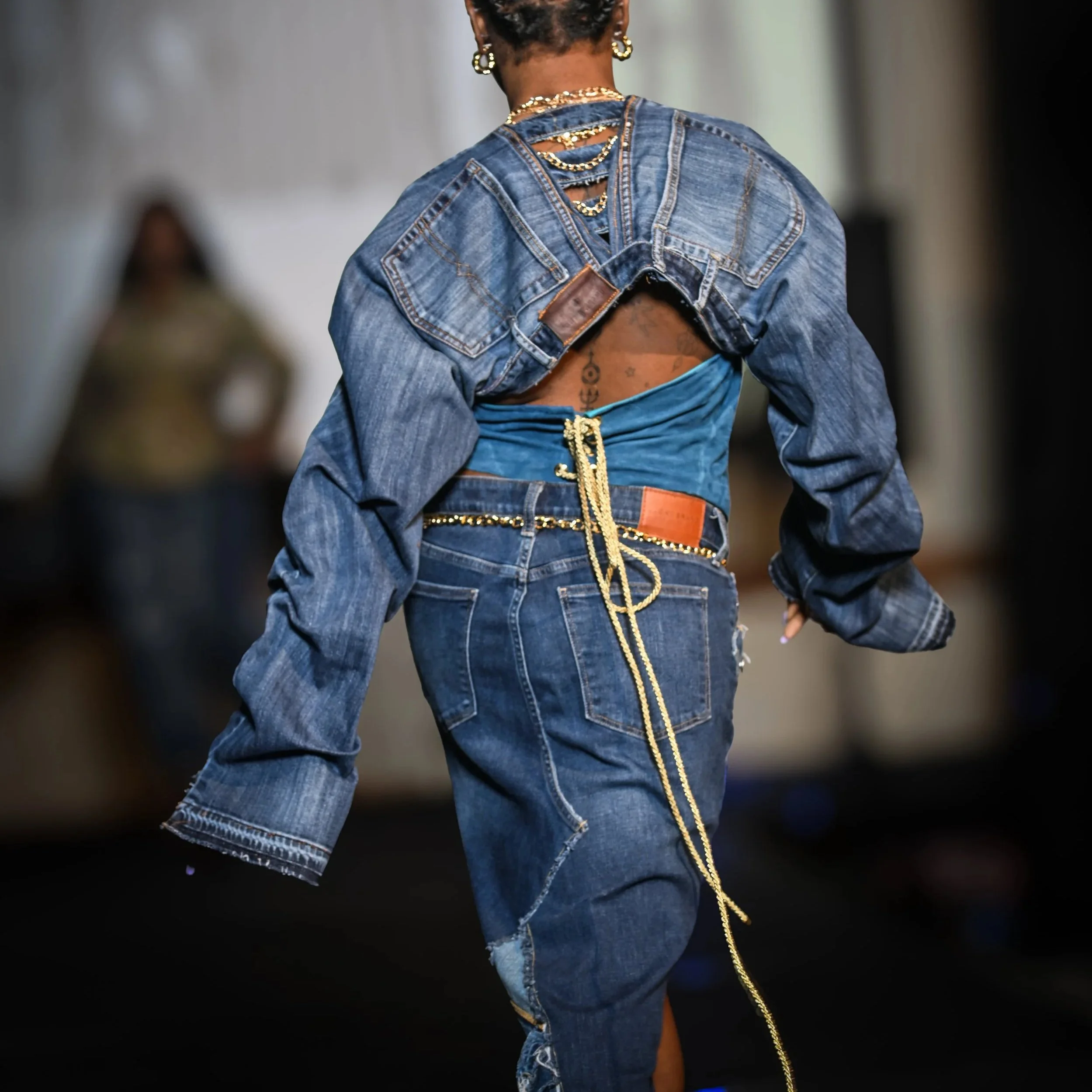 A person walking away from the camera, wearing a handmade custom vintage denim jacket with a custom distressed skirt, a turquoise bandeau top, and layered gold jewelry, including earrings, chains, and a belt with a long gold rope detail.