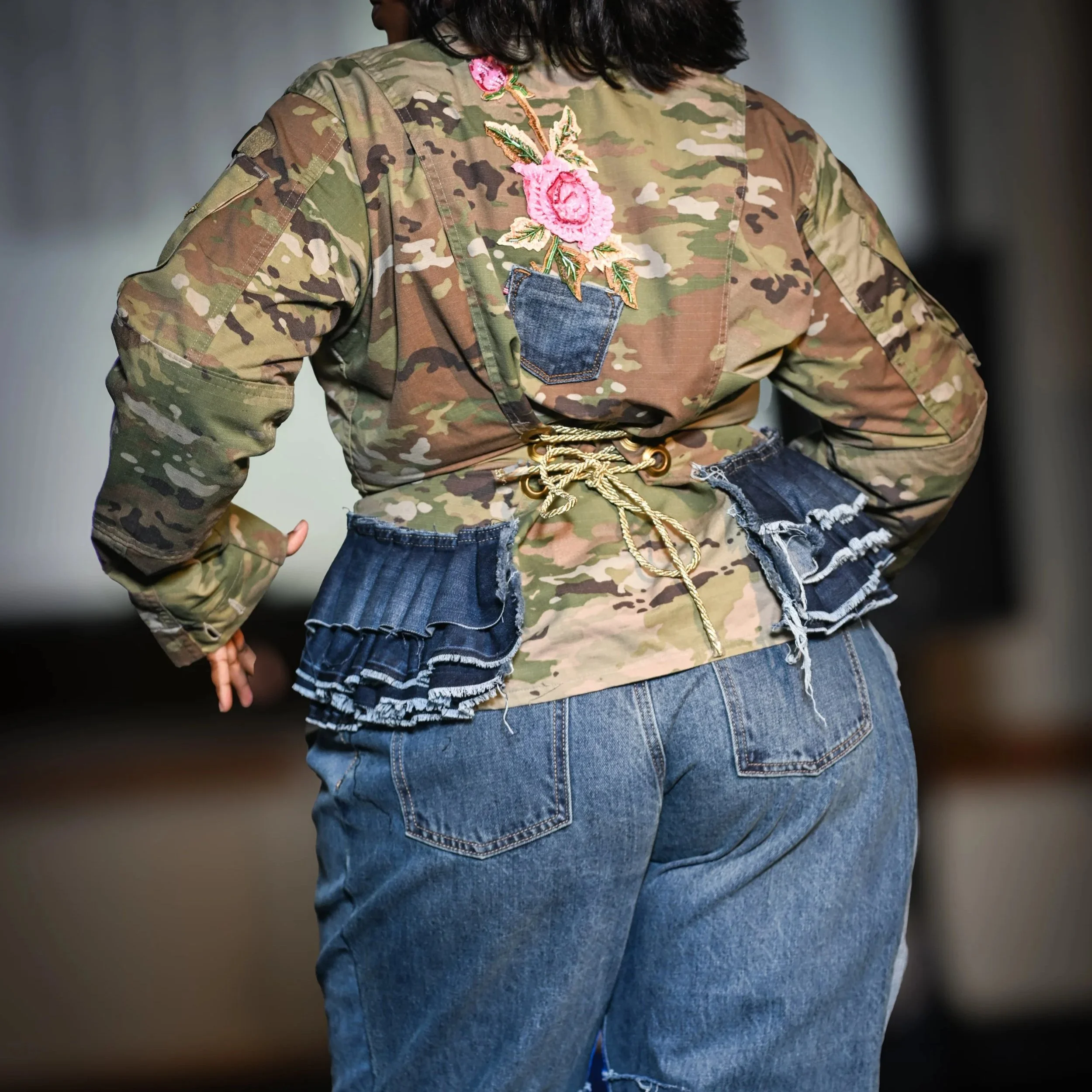 A person wearing a camouflage jacket with pink floral embroidery on the back, paired with frayed denim jeans and a corseted waist, standing in a casual pose.