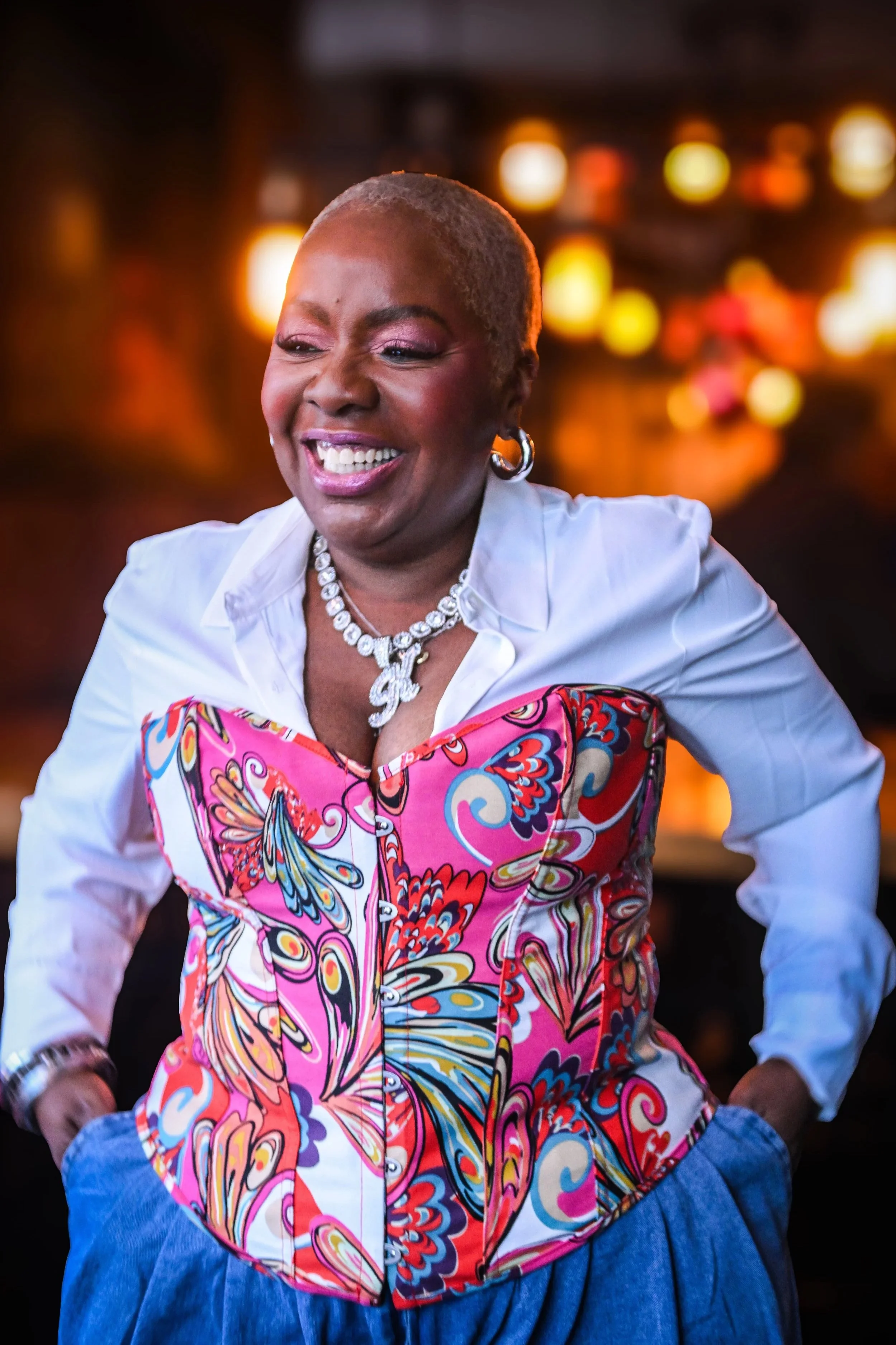 A smiling woman with short gray hair wearing a white shirt and a colorful butterfly-patterned corset, accessorized with a diamond necklace and hoop earrings, standing with her hands in her pockets against a blurred background with warm lights.