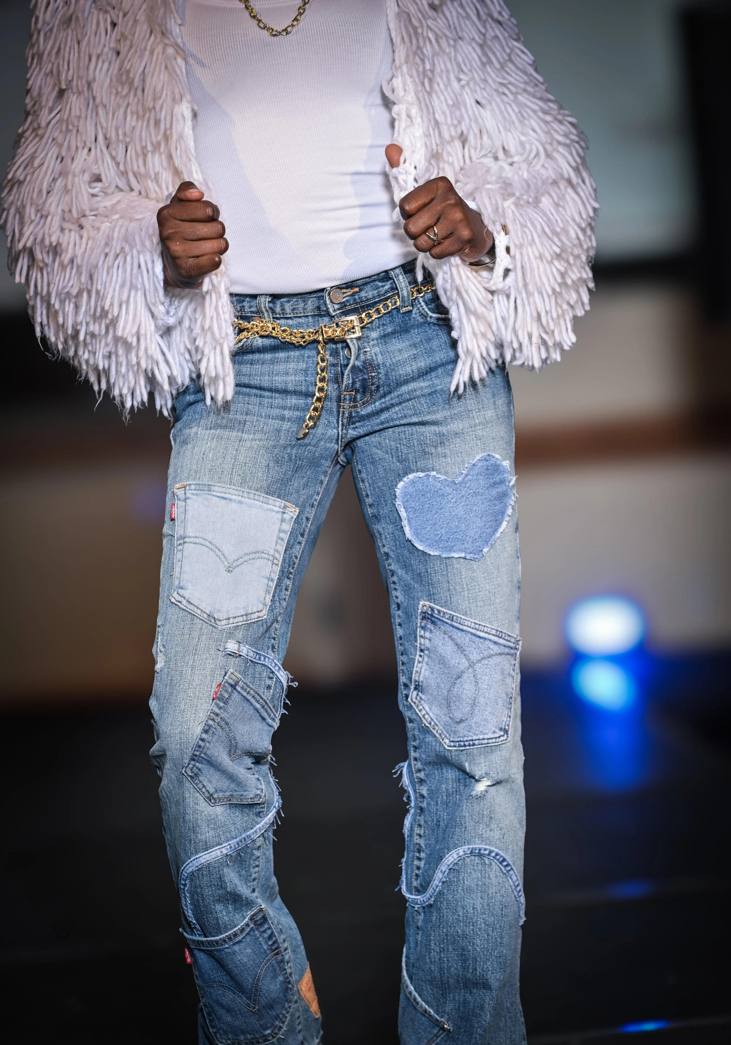 Close-up of a person wearing distressed jeans with heart and pocket patches, a white shirt, crocheted white jacket, gold chain, and rings, with a blurred background.
