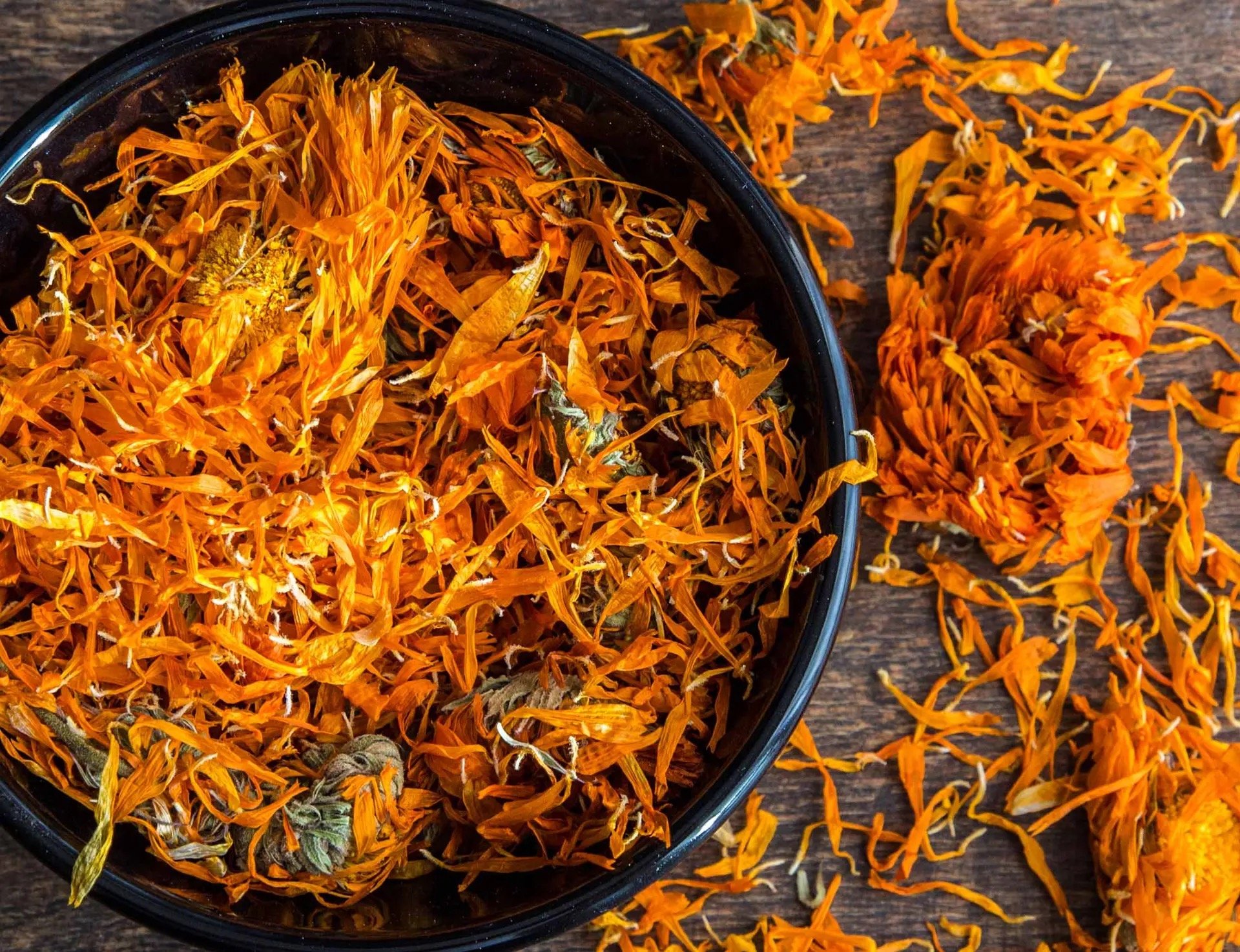 Close-up of dried marigold flowers in a black bowl and scattered on a wooden surface.