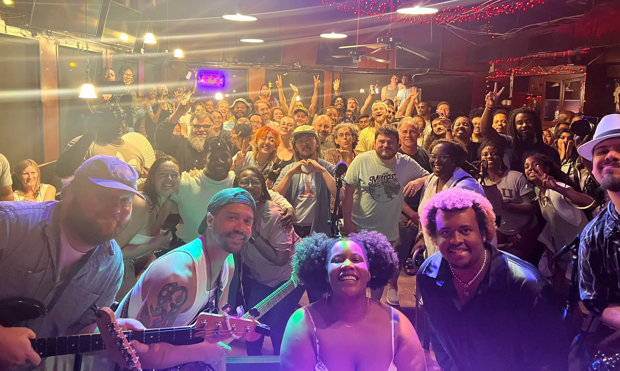 A large group of people inside a bar or club, some holding musical instruments, smiling and posing for a group photo. The setting has colorful lighting and festive decorations.