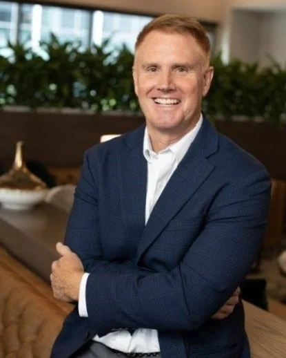A smiling man in a blue suit and white shirt standing indoors with arms crossed, in front of a blurred background with greenery.