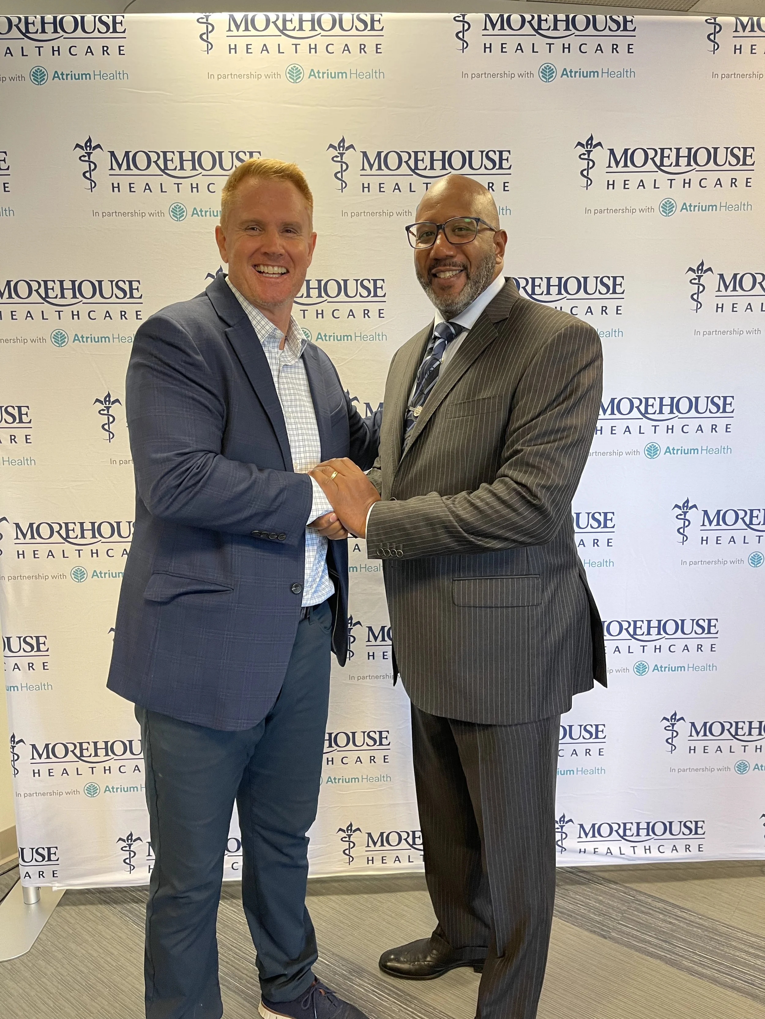 Two men in formal suits shaking hands and smiling in front of a backdrop with 'Morehouse Healthcare' and 'Atrium Health' logos.