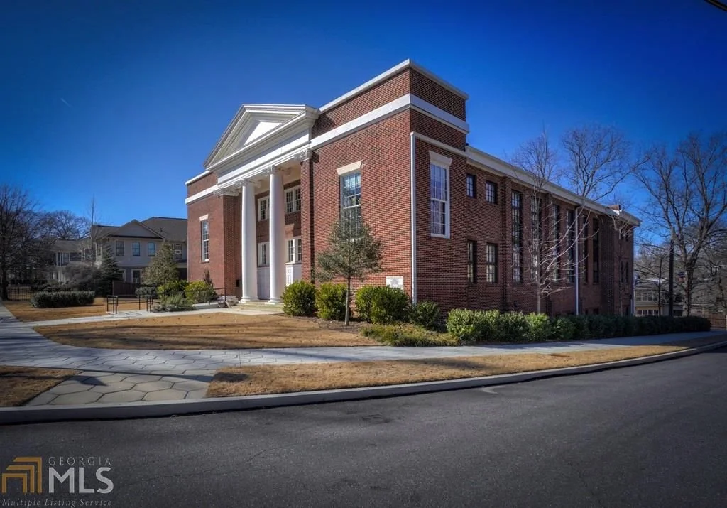 A red brick building with white columns and a triangular pediment; it is set in a landscaped area with a sidewalk, trees, bushes, and a clear blue sky.