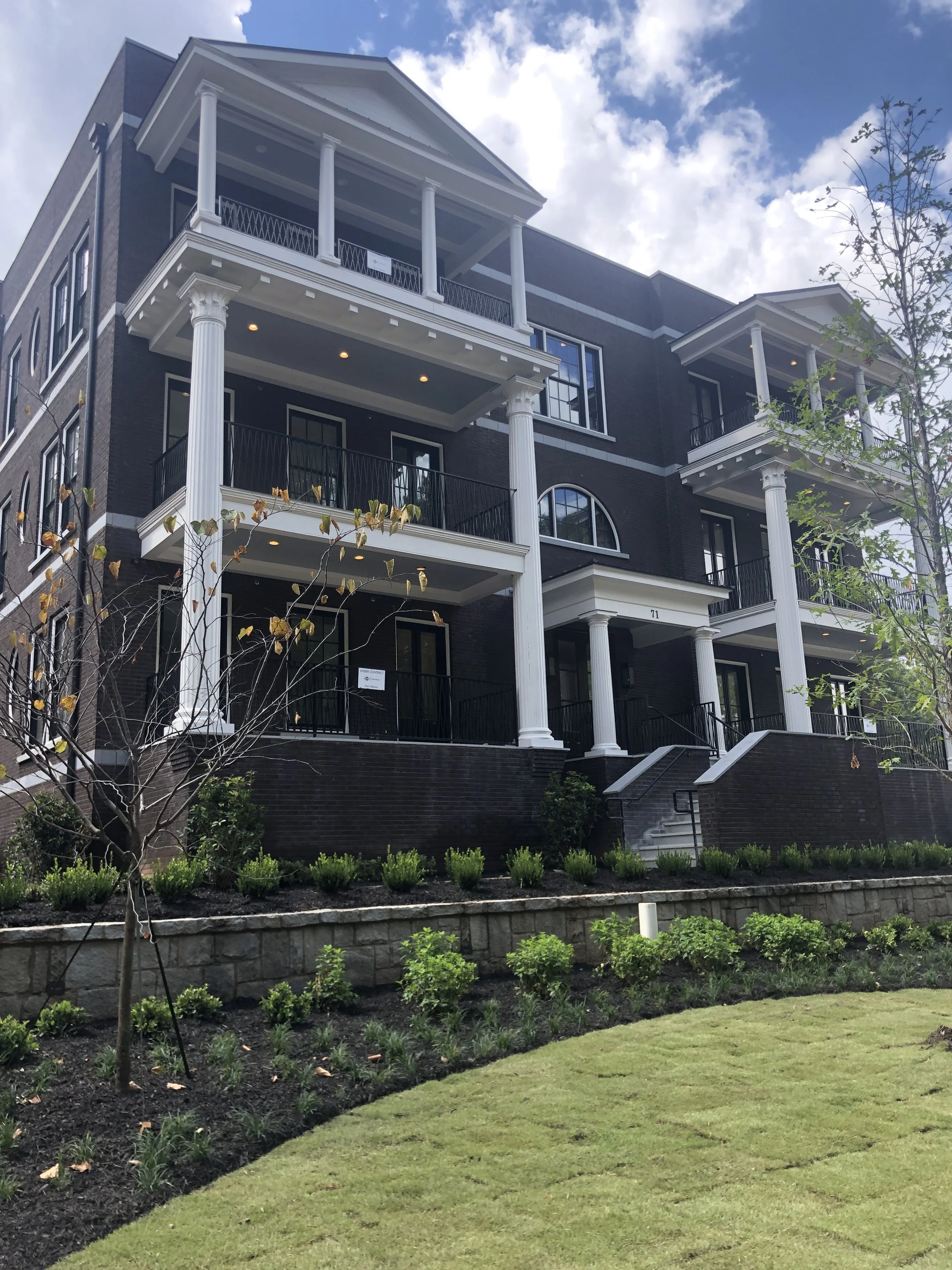 A modern multi-story residential building with black exterior walls and white accents, multiple balconies with black railings, and decorative white columns. The building is surrounded by landscaped greenery and a well-maintained lawn under a partly cloudy sky.