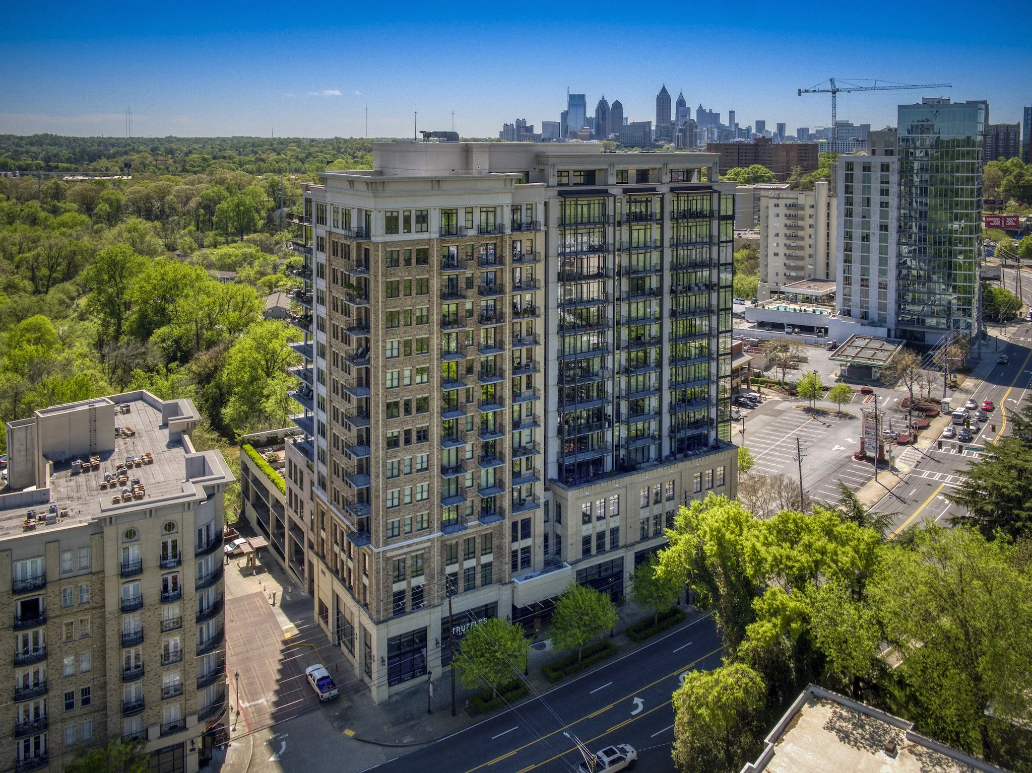 Aerial view of a tall modern apartment building situated next to a green park and across from a parking lot, with a city skyline in the background under a clear blue sky.