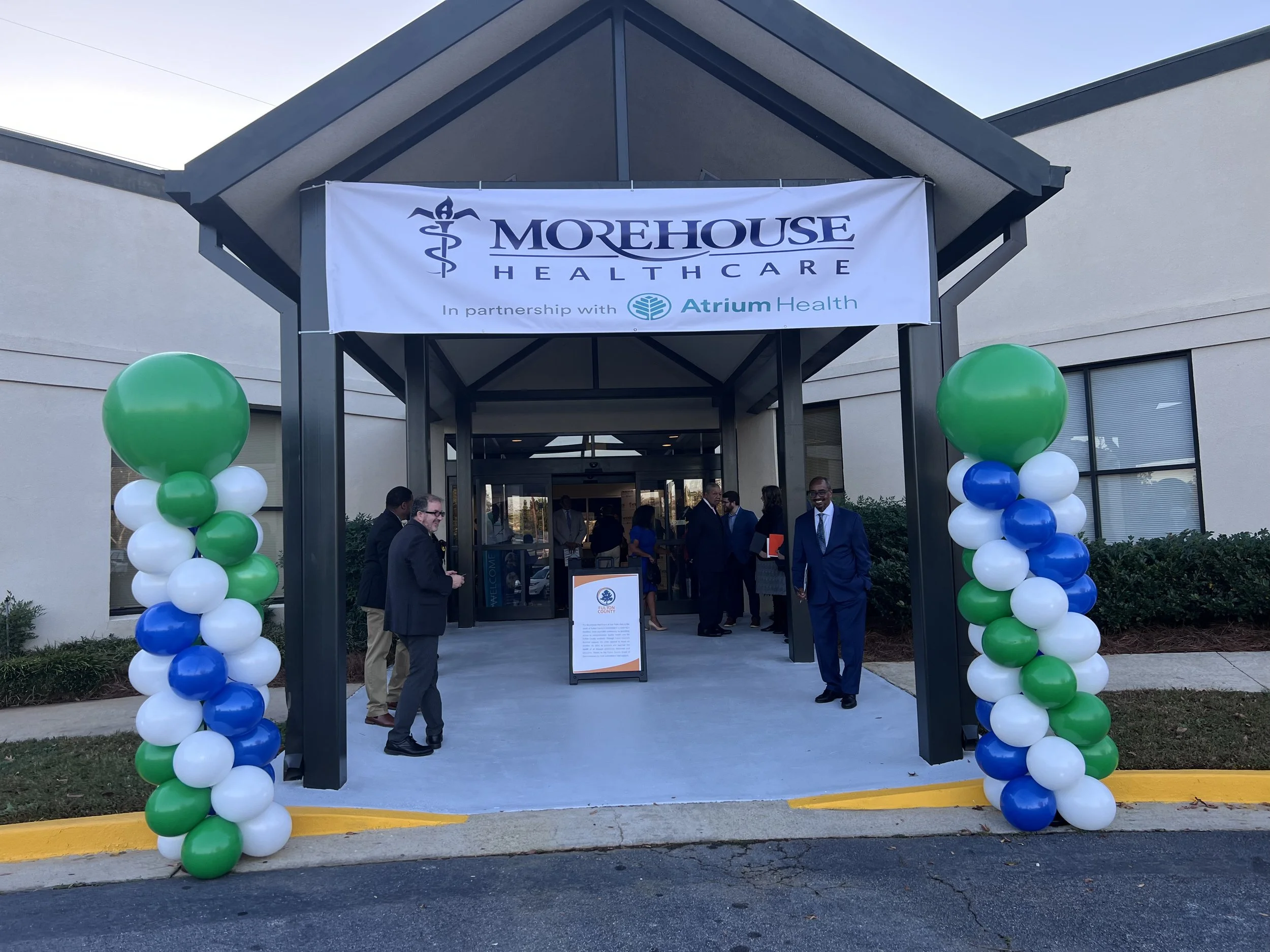 Entrance to Morehouse Healthcare building decorated with green, white, blue balloons on either side. A banner above reads 'MOREHOUSE HEALTHCARE' in partnership with Atrium Health. People are gathered outside, some dressed formally.