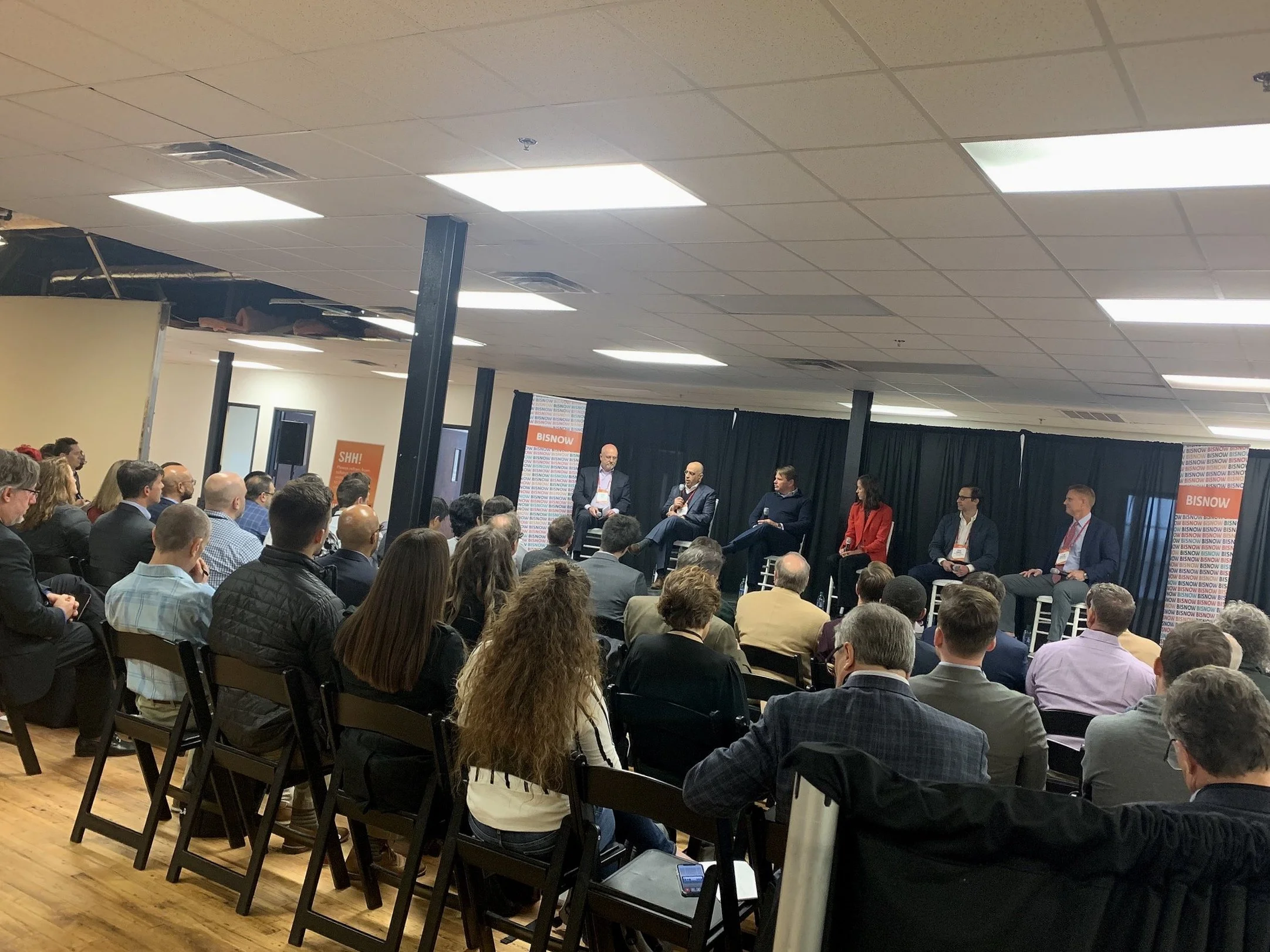 A panel discussion at a conference, with six people on stage and a large audience seated facing the stage. The setting appears to be indoors with ceiling lights and black curtains at the back of the stage. There are informational banners on either side of the stage.