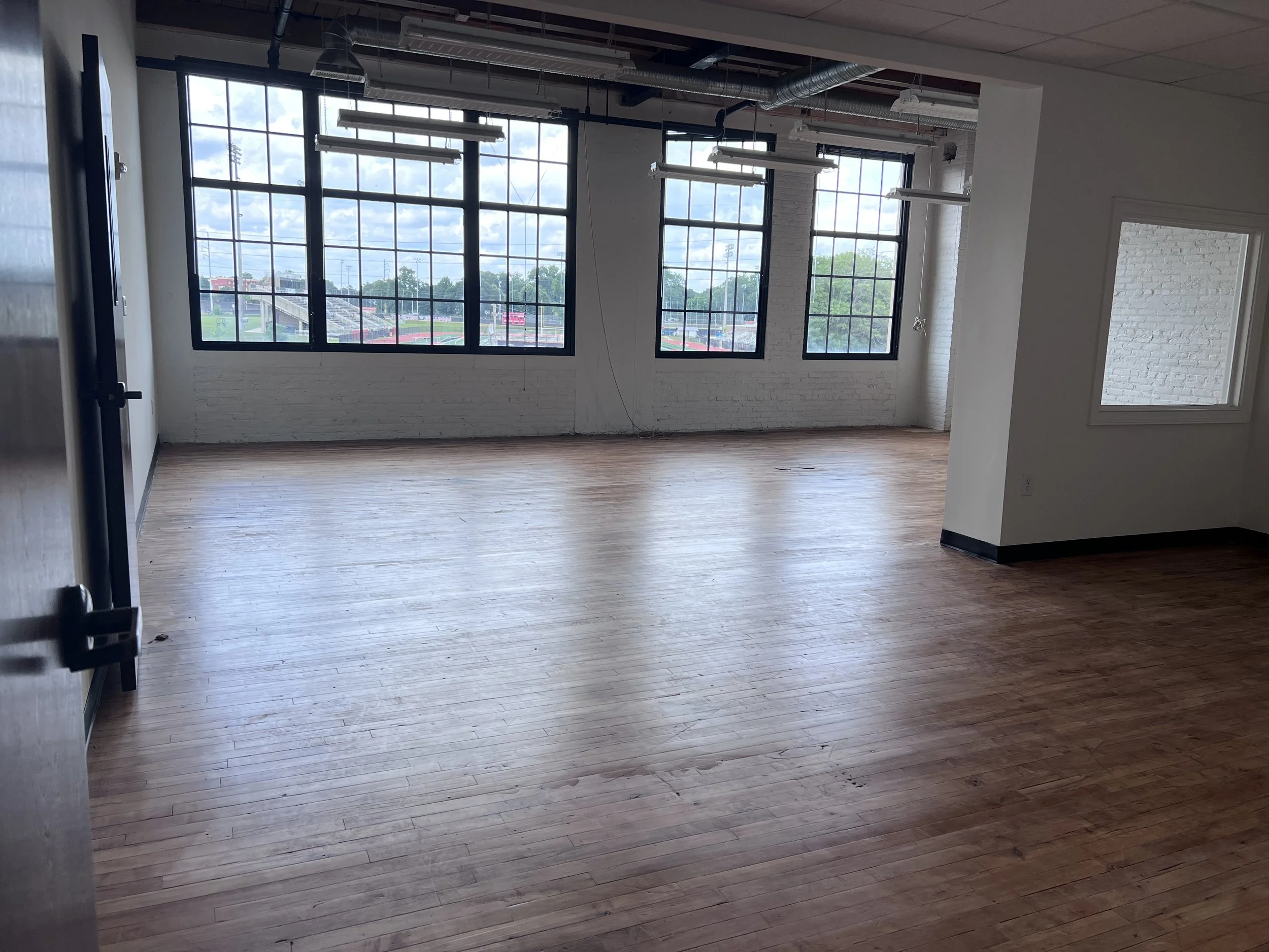 Empty room with large windows, hardwood floors, exposed ceiling pipes, and white brick walls, natural light coming in.