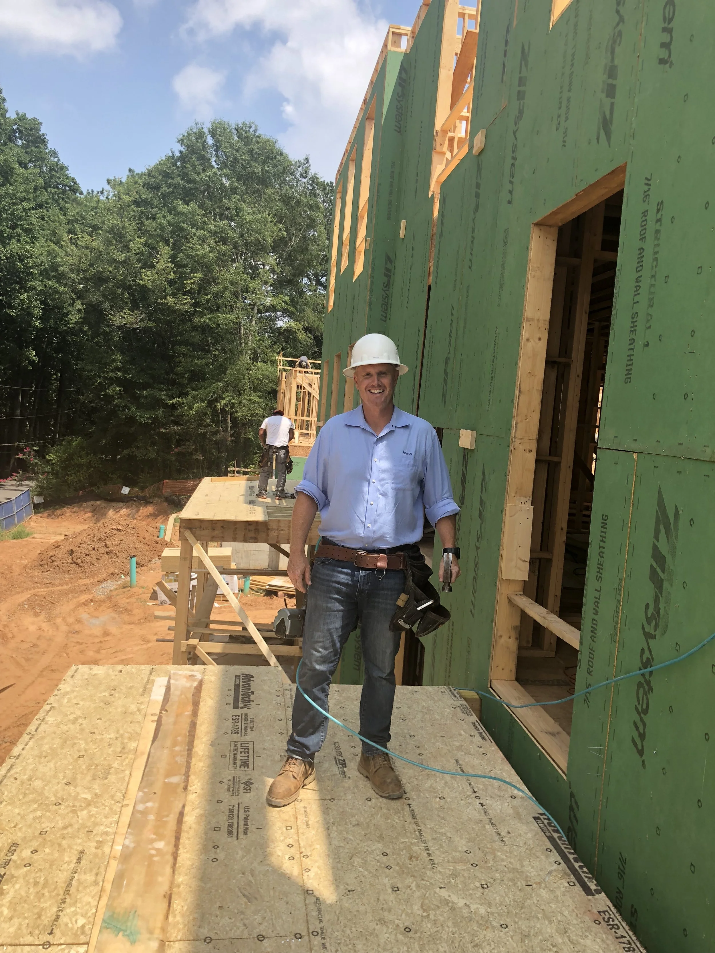 A construction worker wearing a white hard hat and blue shirt standing on a wooden platform at a building site under a partly cloudy sky. Two other workers are visible working on the building in the background.
