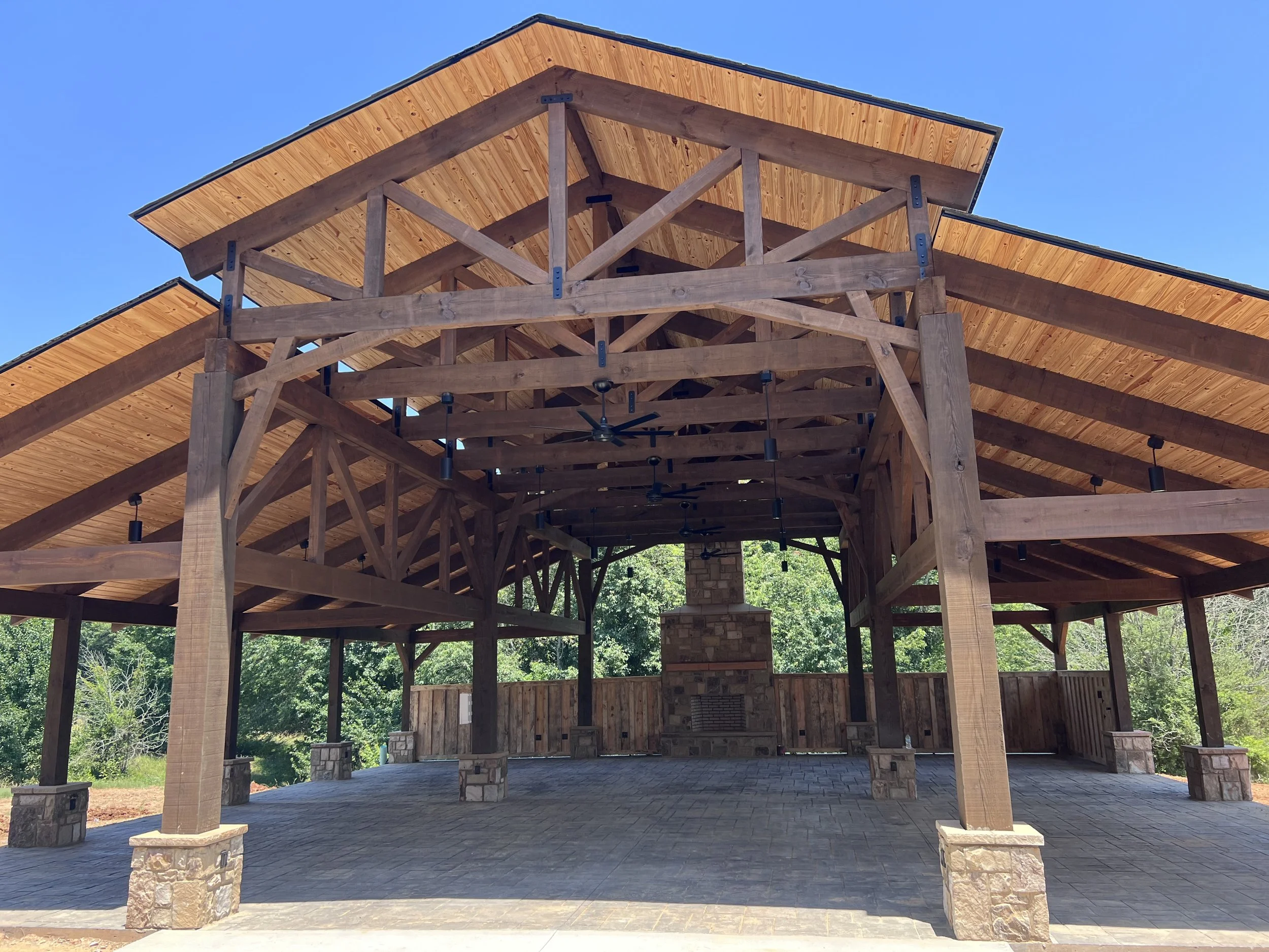 An outdoor wooden pavilion with a chimney, ceiling fans, and stone support columns, set against a clear blue sky and surrounded by green trees.