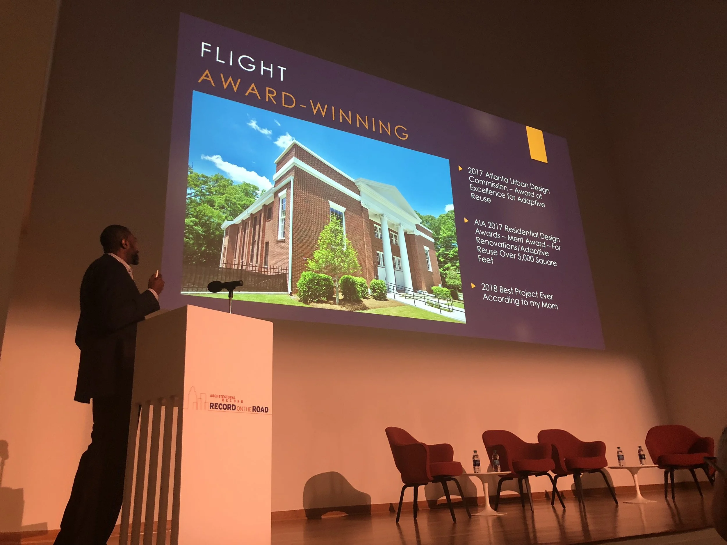 A man in a suit giving a presentation on stage with a large screen behind him. The screen displays a building and a list of awards and recognitions related to architecture and design.