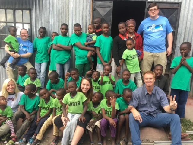 Group photo of children and young adults, some wearing green t-shirts with 'SANTA' printed on them, standing and sitting outside a rustic building with corrugated metal walls, alongside a few adults.
