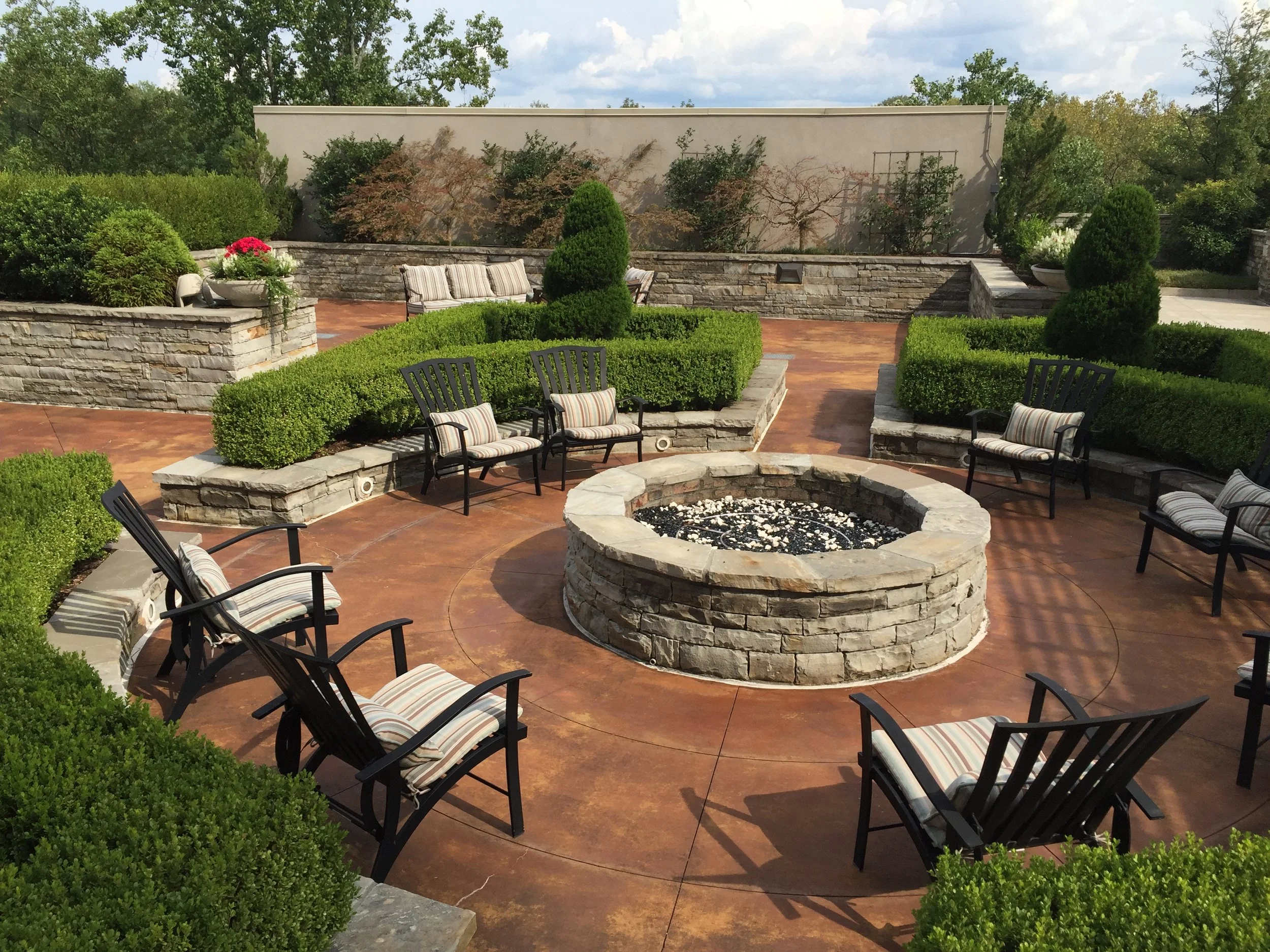Outdoor patio with a circular fire pit at center, surrounded by black chairs with striped cushions, green bushes, and a low stone wall with additional seating and flower pots.