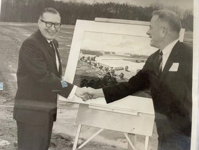 Two men in suits shaking hands outdoors, with a large landscape painting on an easel between them. The landscape depicts fields and trees, suggesting a rural or farmland setting.