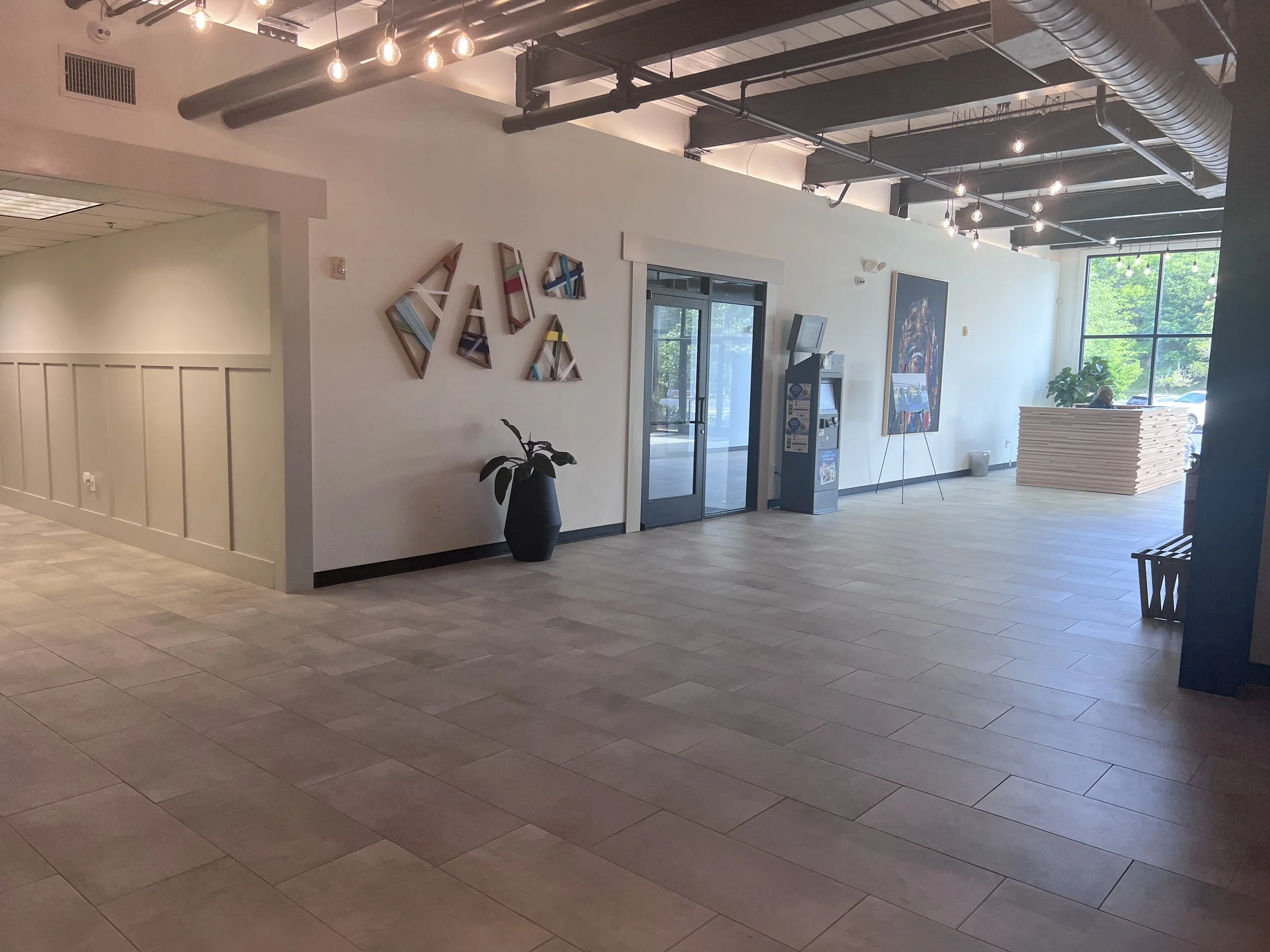 Modern office lobby with large window, artwork on white walls, potted plant, and seating area.