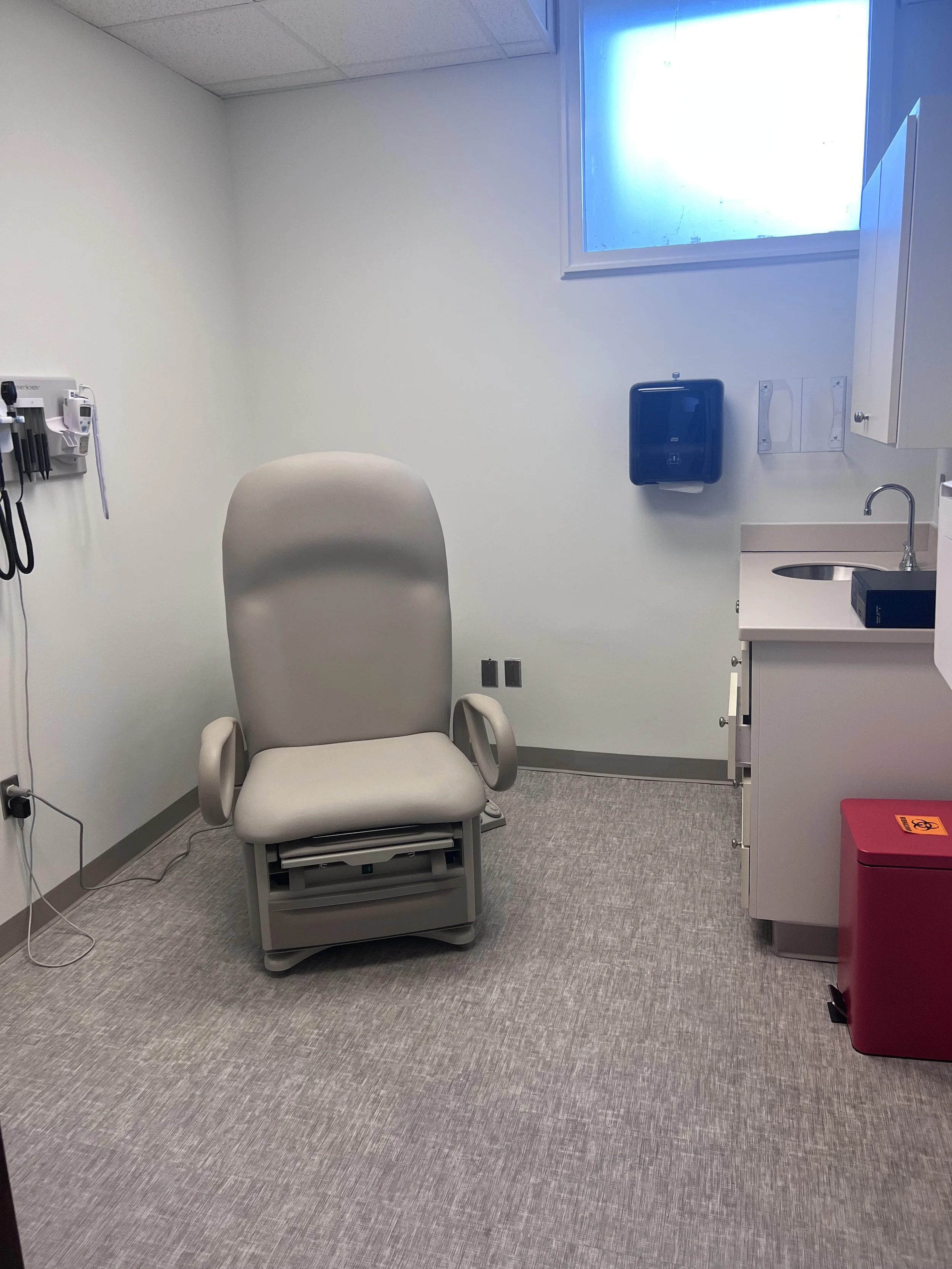 Empty medical examination room with a beige chair, medical instruments on the wall, a sink, a red biohazard waste container, and a window with frosted blue glass.