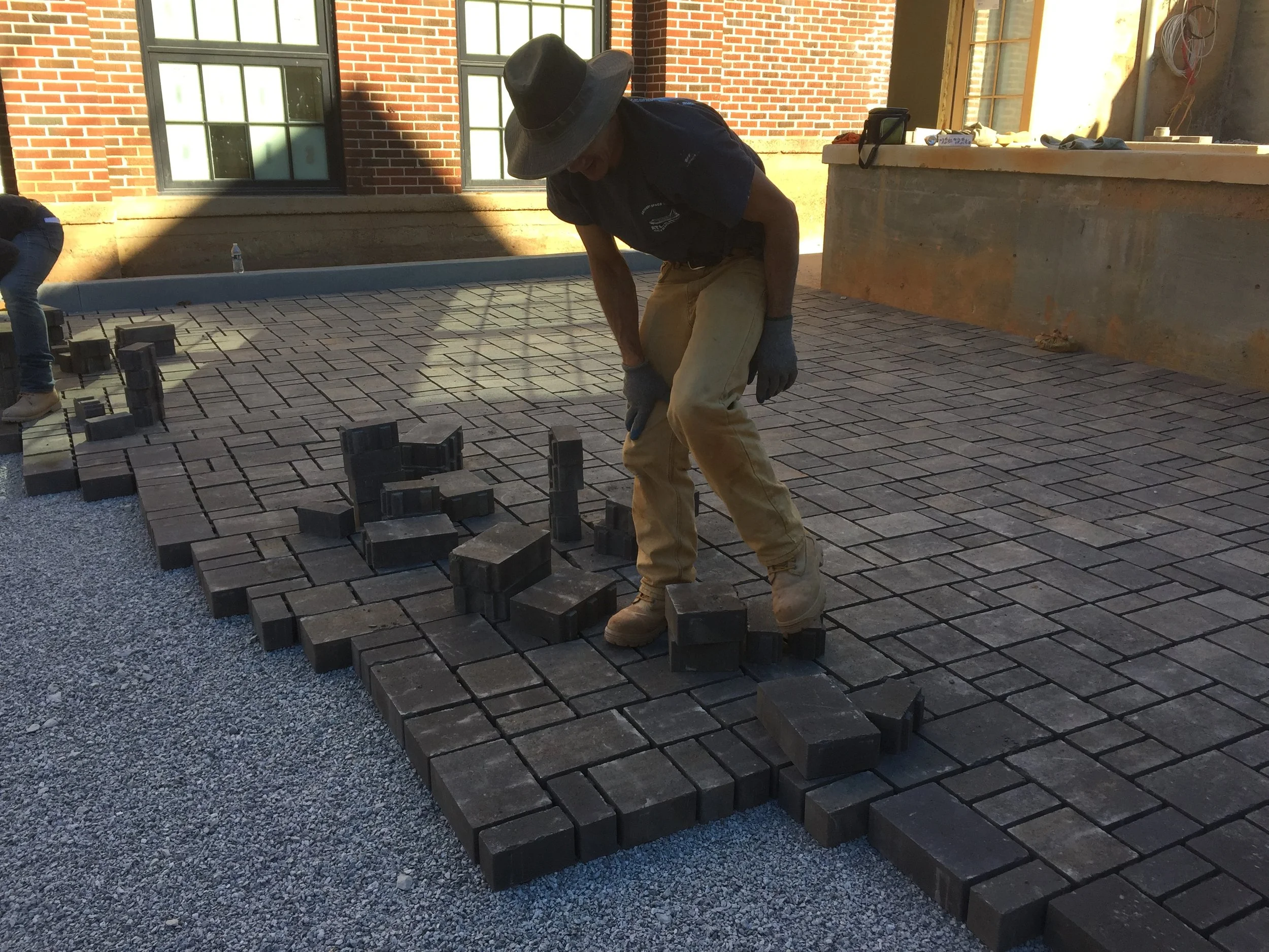 A construction worker installing gray bricks on a patio or walkway, bending down with gloves on, wearing a wide-brimmed hat, dark shirt, and khaki pants. Other workers and building materials are visible in the background.