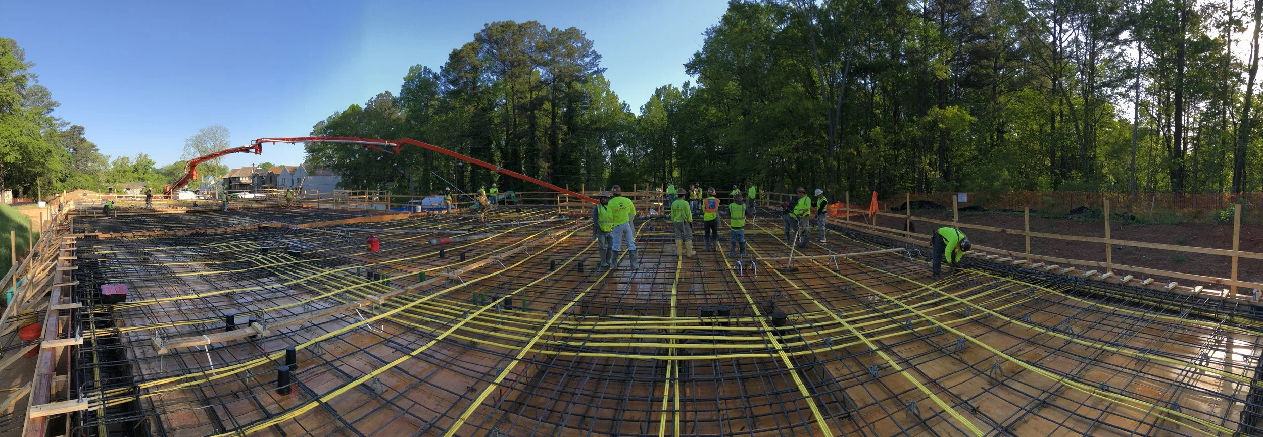 Construction workers installing rebar and electrical conduits for a building foundation in a wooded area during daytime.