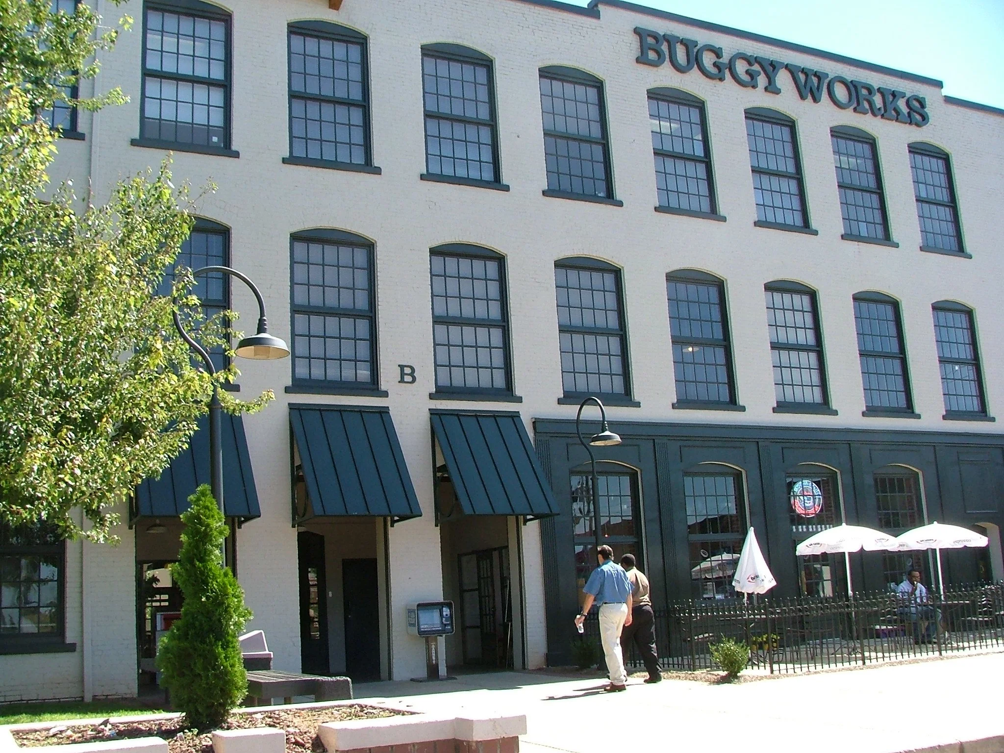 A large multi-story building with the sign "BUGGY WORKS" at the top. The building has large windows and black awnings. There are people walking outside and sitting under umbrellas at an outdoor seating area.