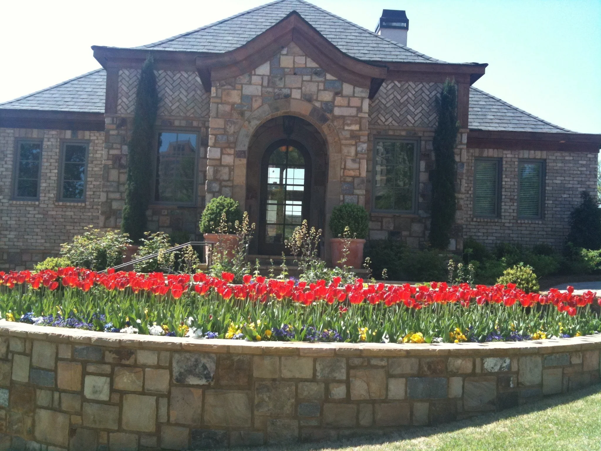 A large house with a stone and brick exterior, multiple windows, and a arched front door, surrounded by a garden with red tulips, potted plants, and trees in pots.