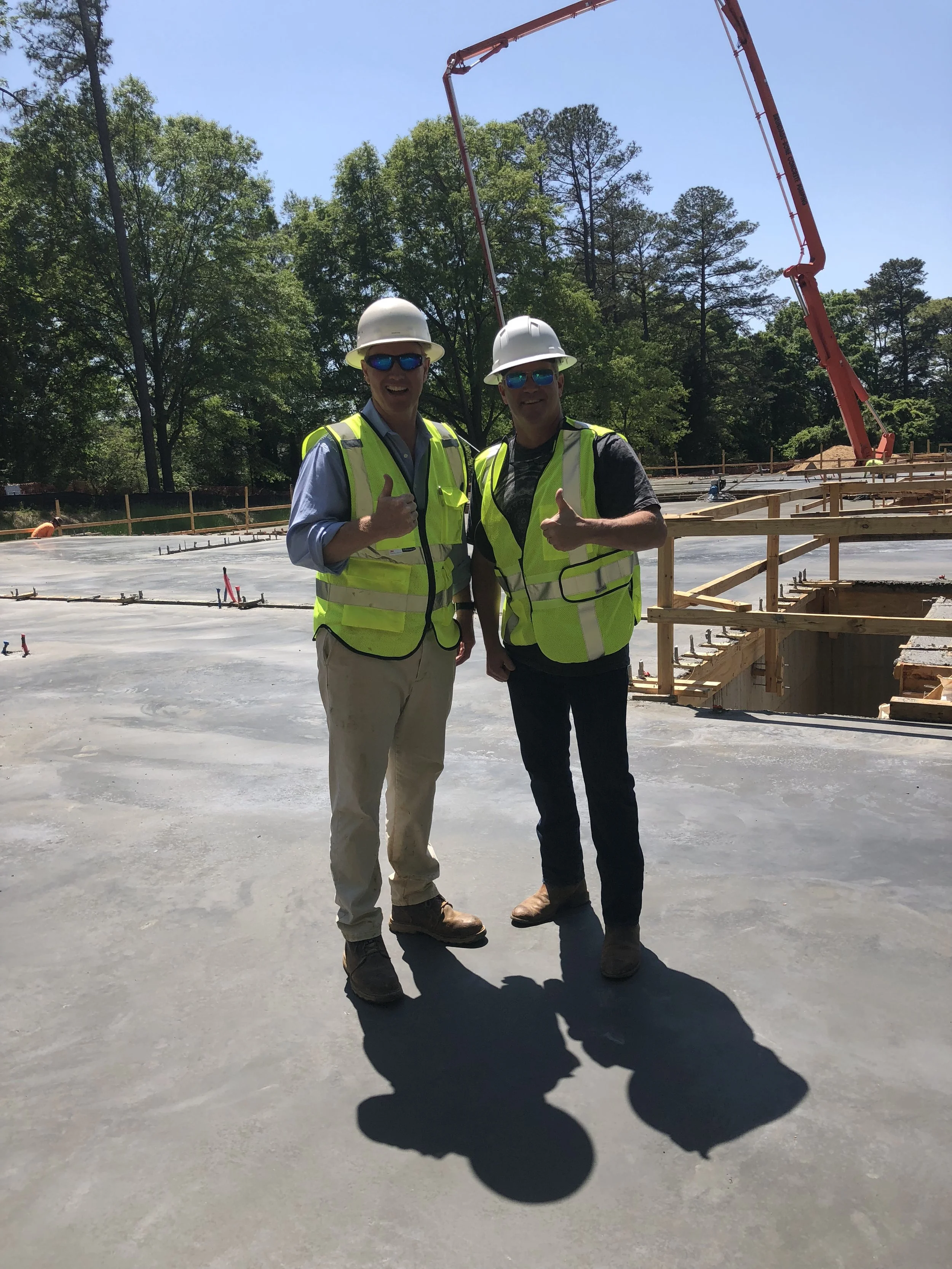 Two construction workers in safety vests and hard hats standing on freshly poured concrete at a construction site, with a crane and trees in the background.
