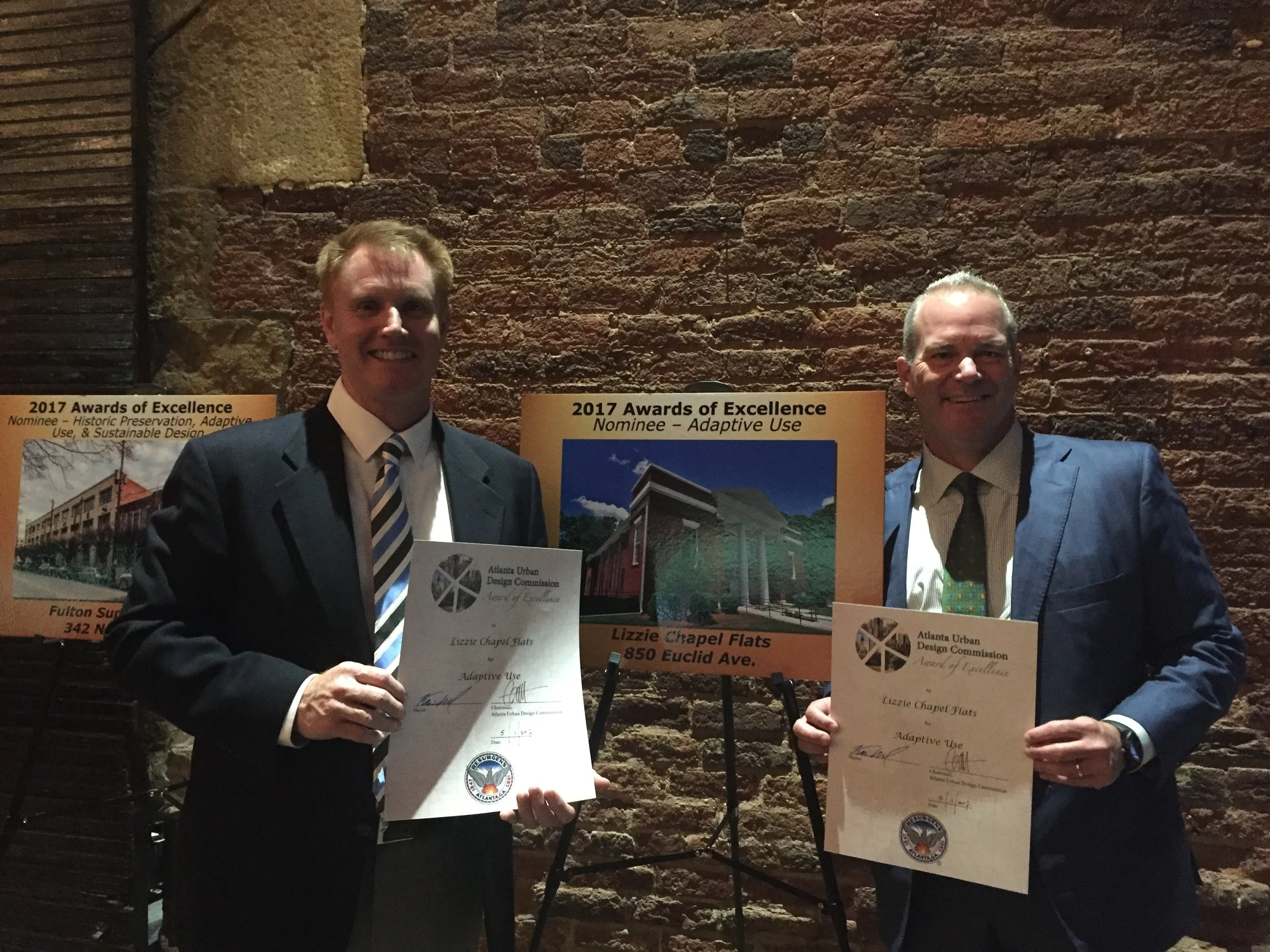 Two men in suits holding awards at the 2017 Awards of Excellence ceremony, standing in front of posters and a brick wall.