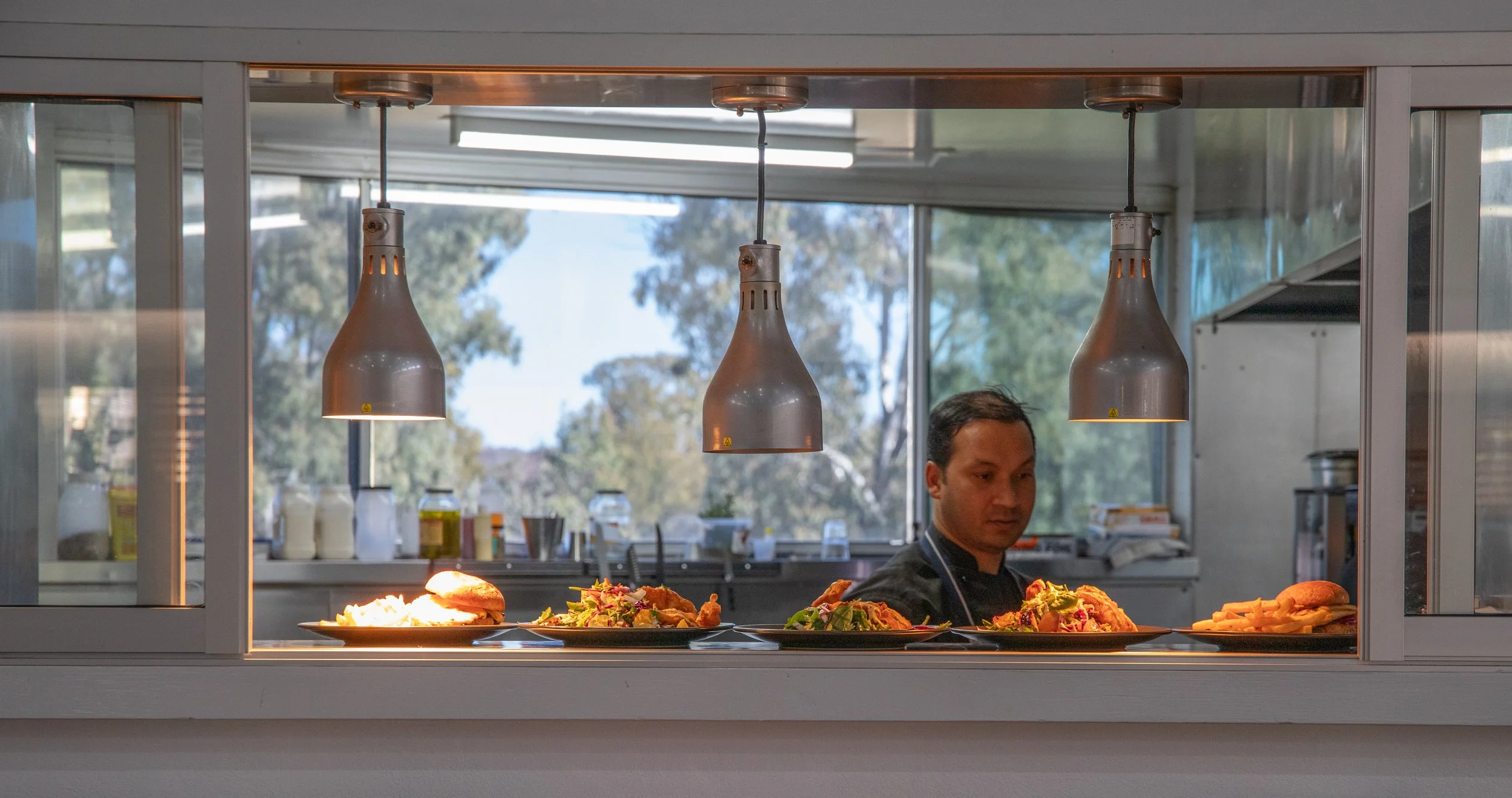 A man working in a restaurant kitchen seen through a glass window with three pendant lights hanging above plates of food, including sandwiches and salads, on a counter.