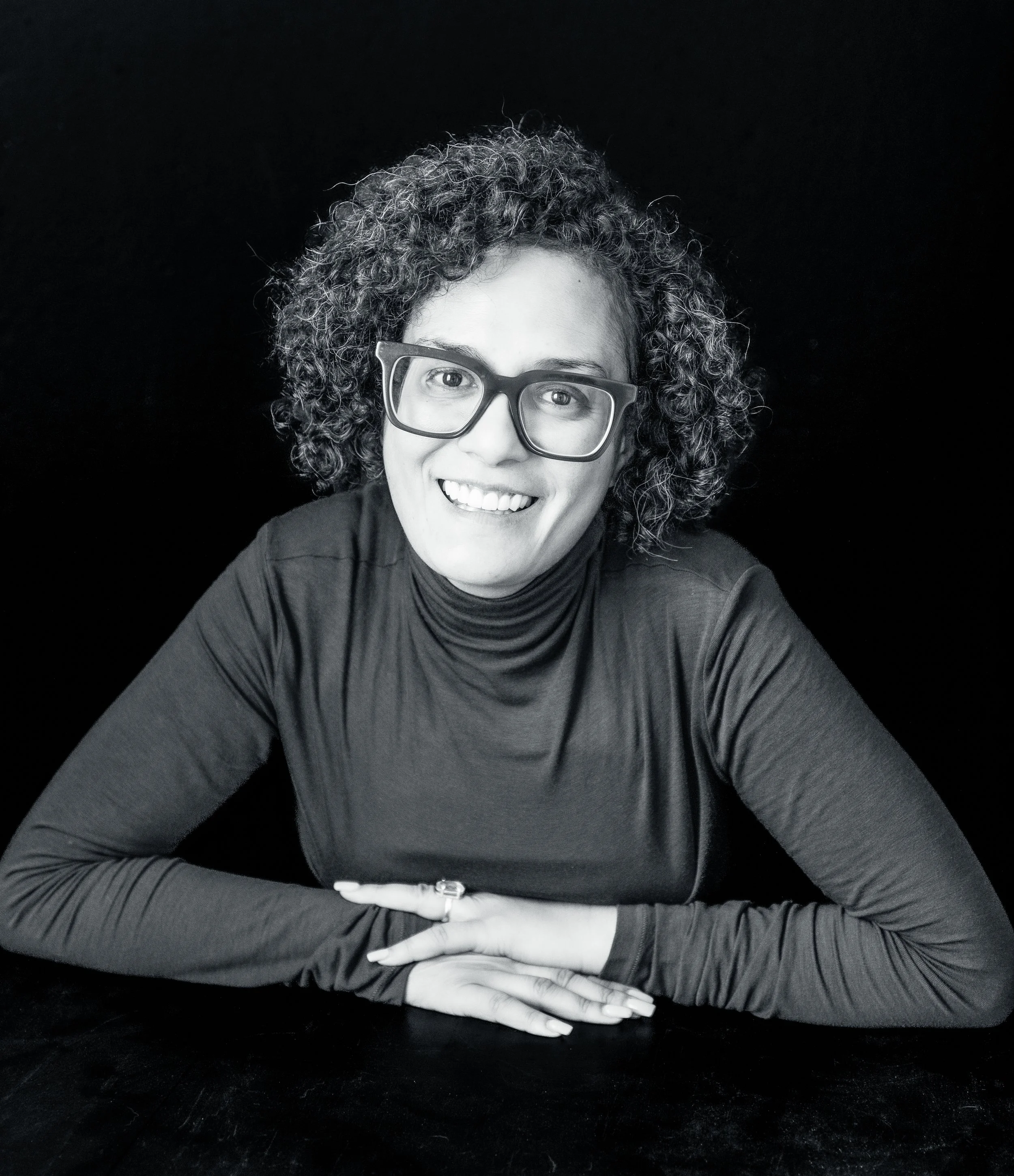 A woman with curly hair, glasses, and a turtleneck top sitting at a dark surface against a black background, smiling at the camera.
