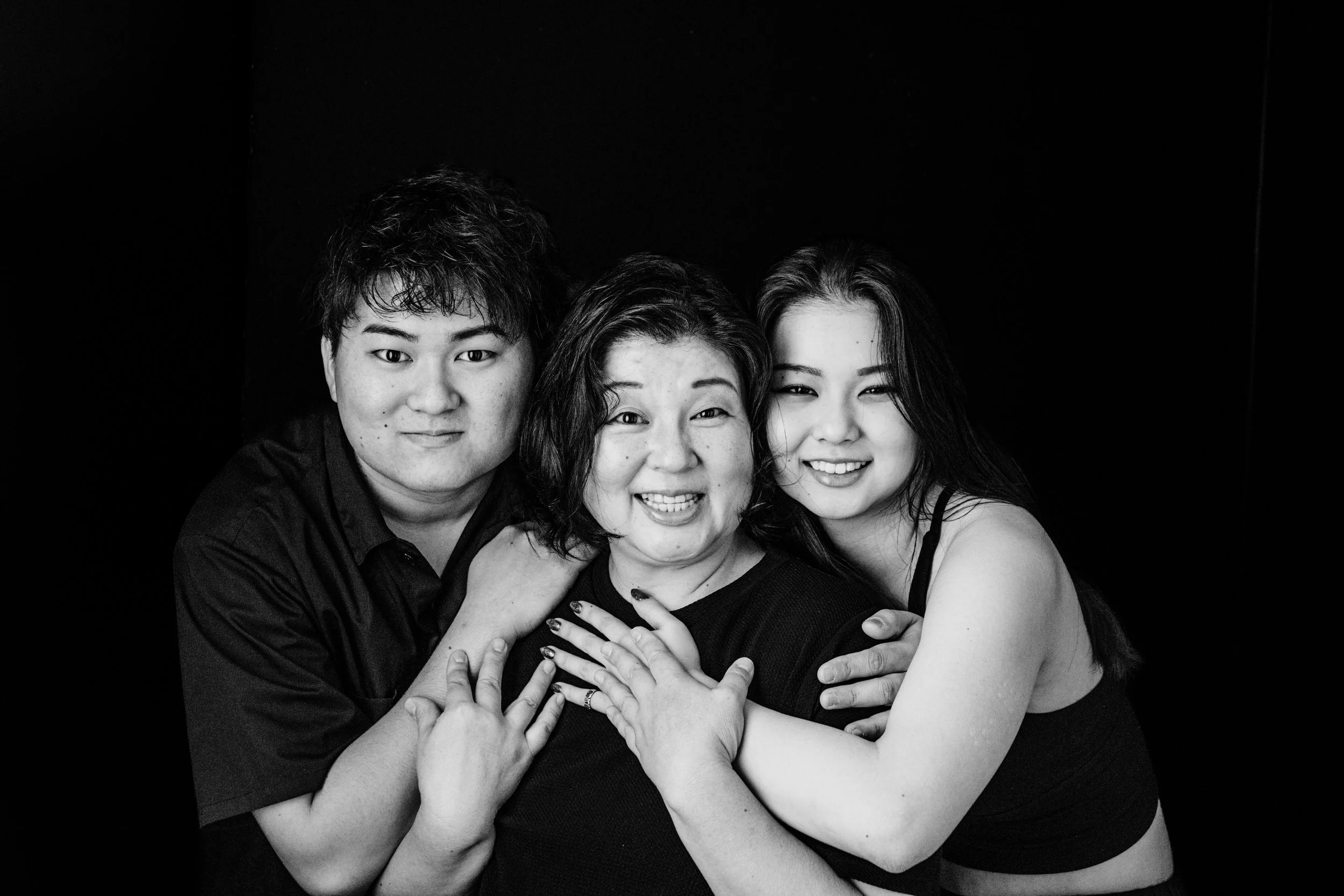 Black and white photo of three smiling women embracing each other against a dark background.