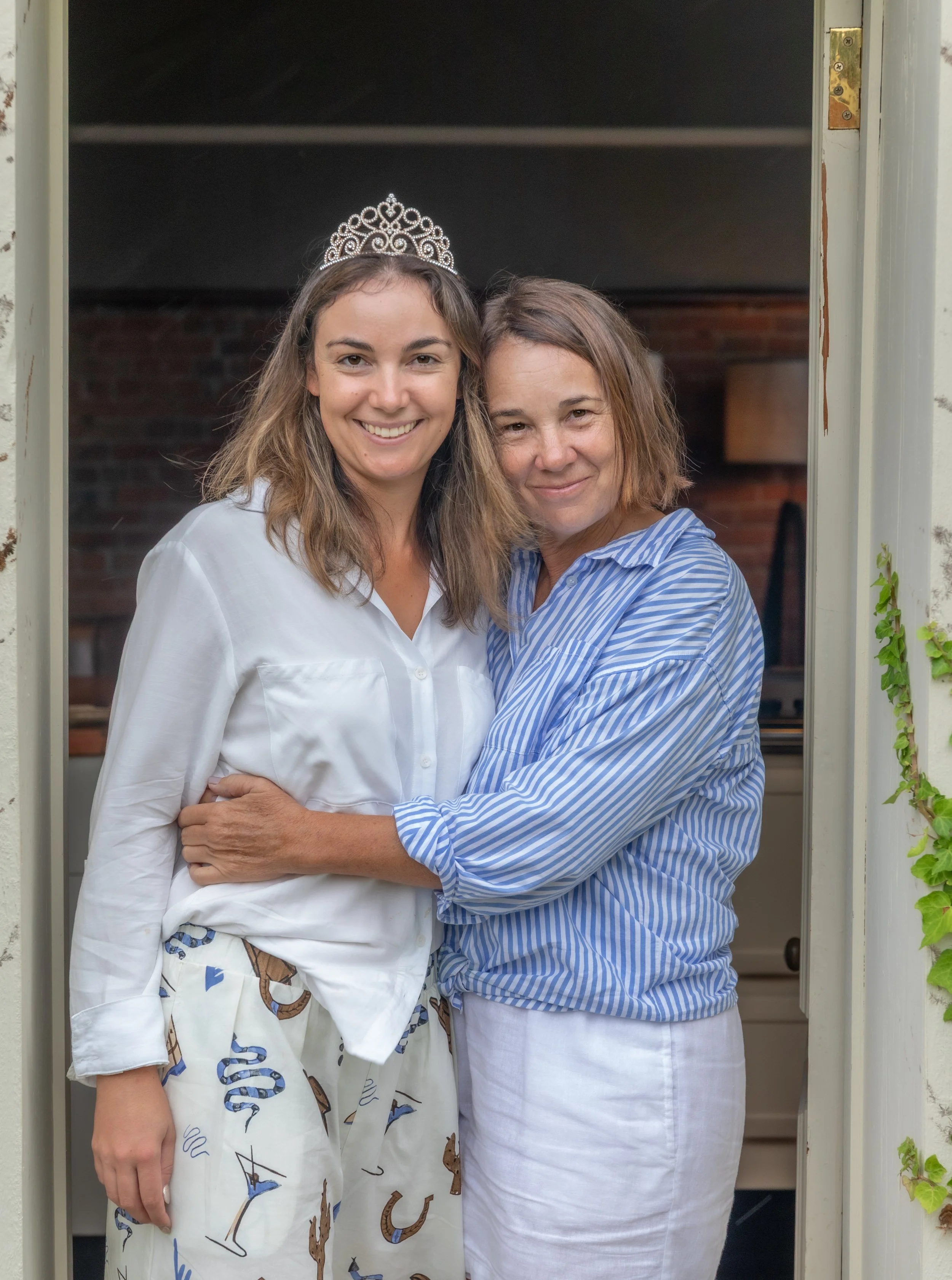 Two women smiling and hugging each other in a doorway. The woman on the left is wearing a white shirt and pants with a nautical print, and a tiara. The woman on the right is wearing a blue and white striped shirt and white pants.