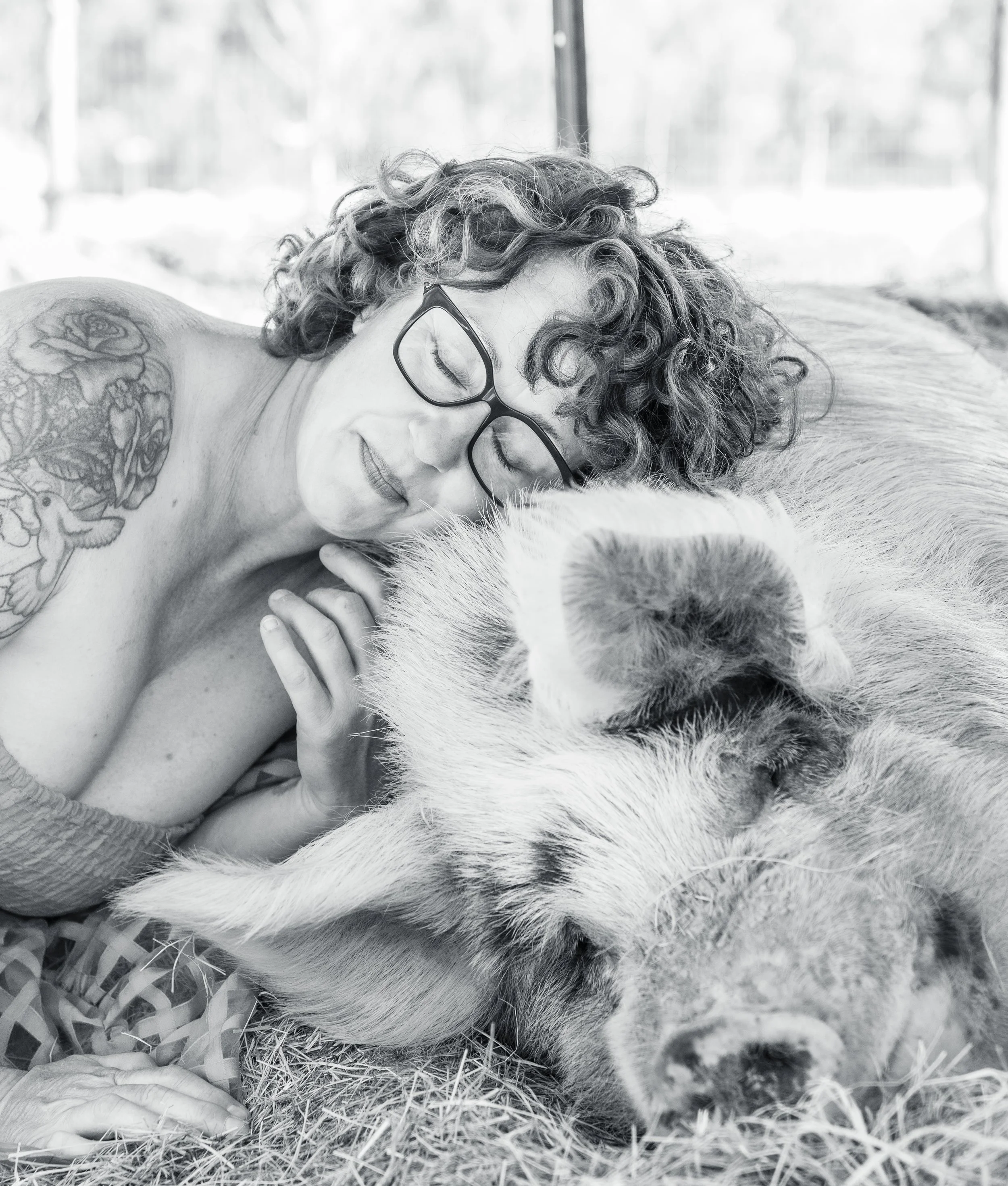 A woman with curly hair and glasses resting her head on a large pig, both lying on straw, in a barn or farm setting.