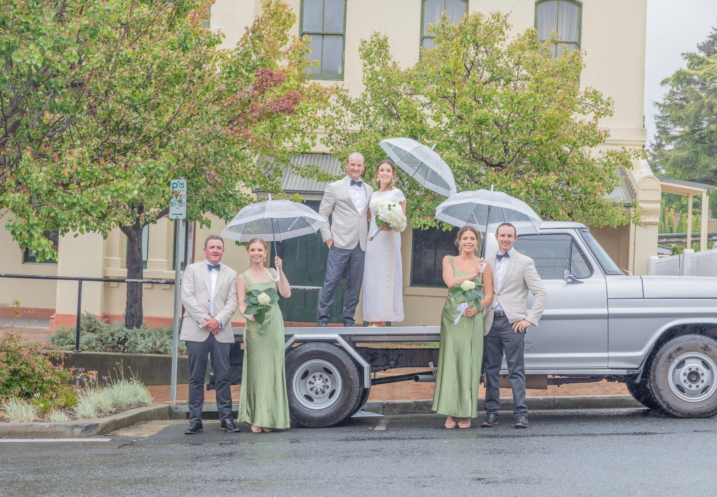 Wedding party on a pickup truck with umbrellas in a rainy outdoor setting. The bride and groom stand in the truck bed, with bridesmaids and groomsmen around them, some holding bouquets and umbrellas. The background features trees and a building.