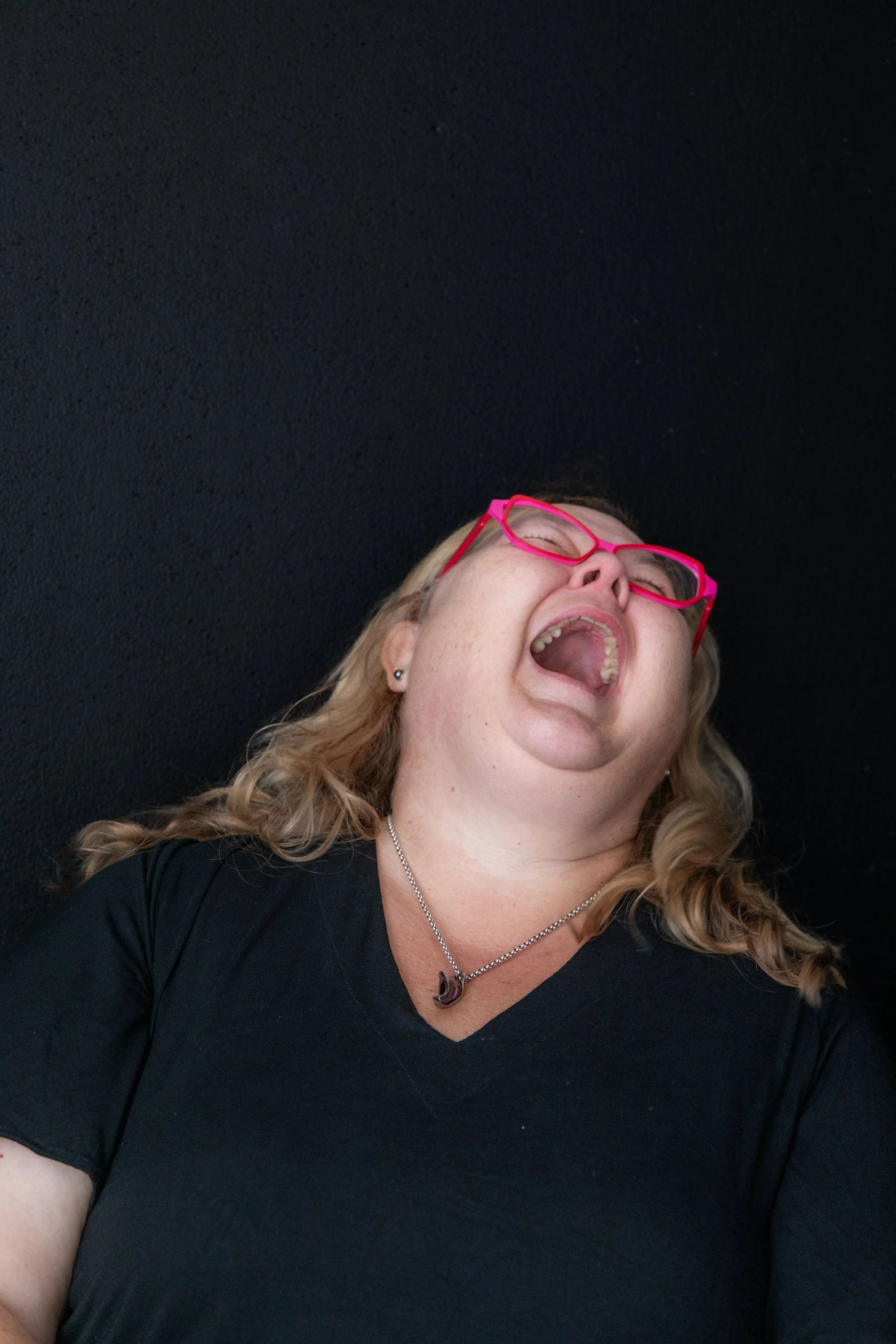 Woman with blonde hair, wearing pink glasses, black shirt, and a necklace; her mouth wide open and head tilted back against a black background.