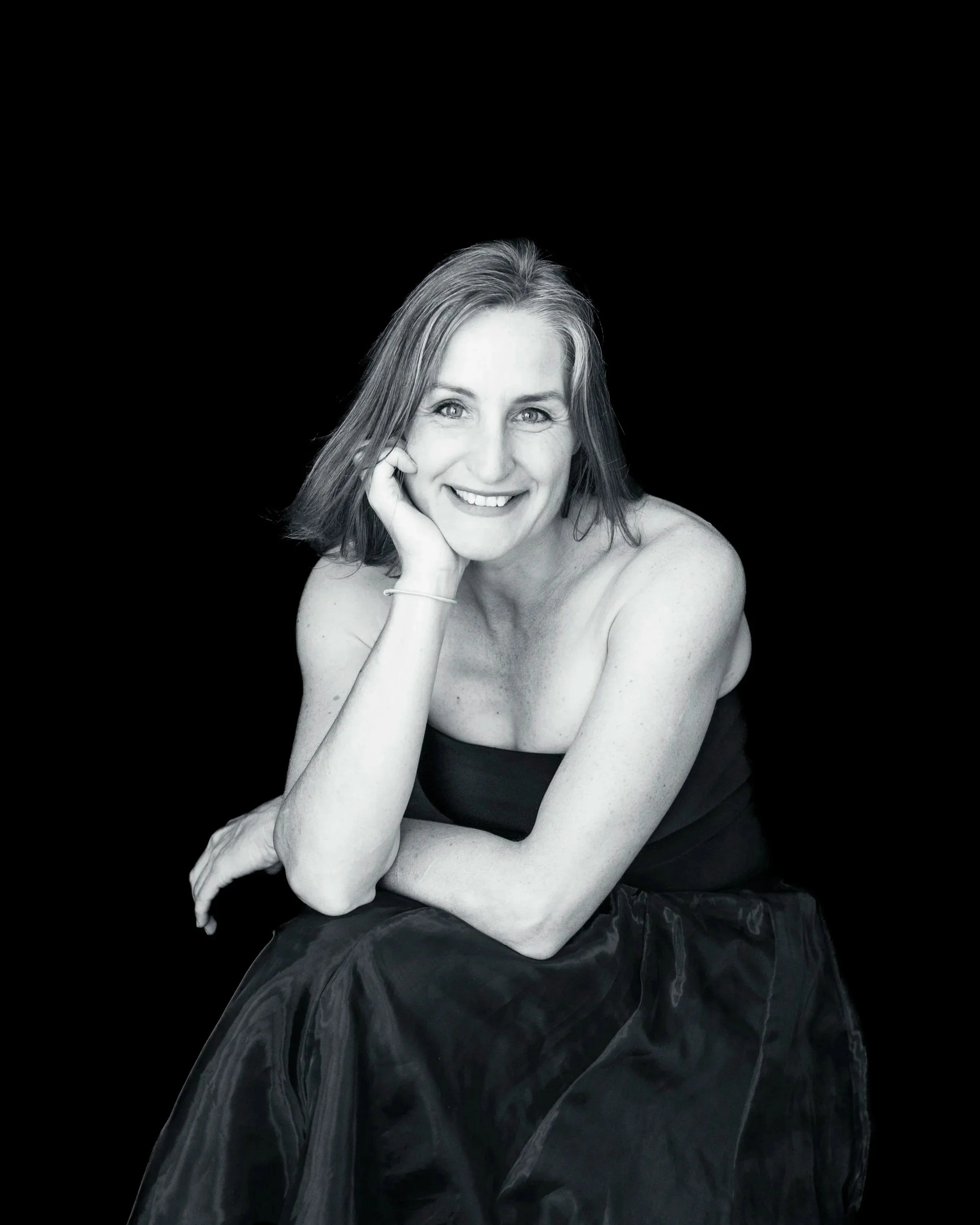 Black and white portrait of a smiling woman with shoulder-length hair, sitting with her chin resting on her hand, wearing a strapless top and a skirt against a black background.