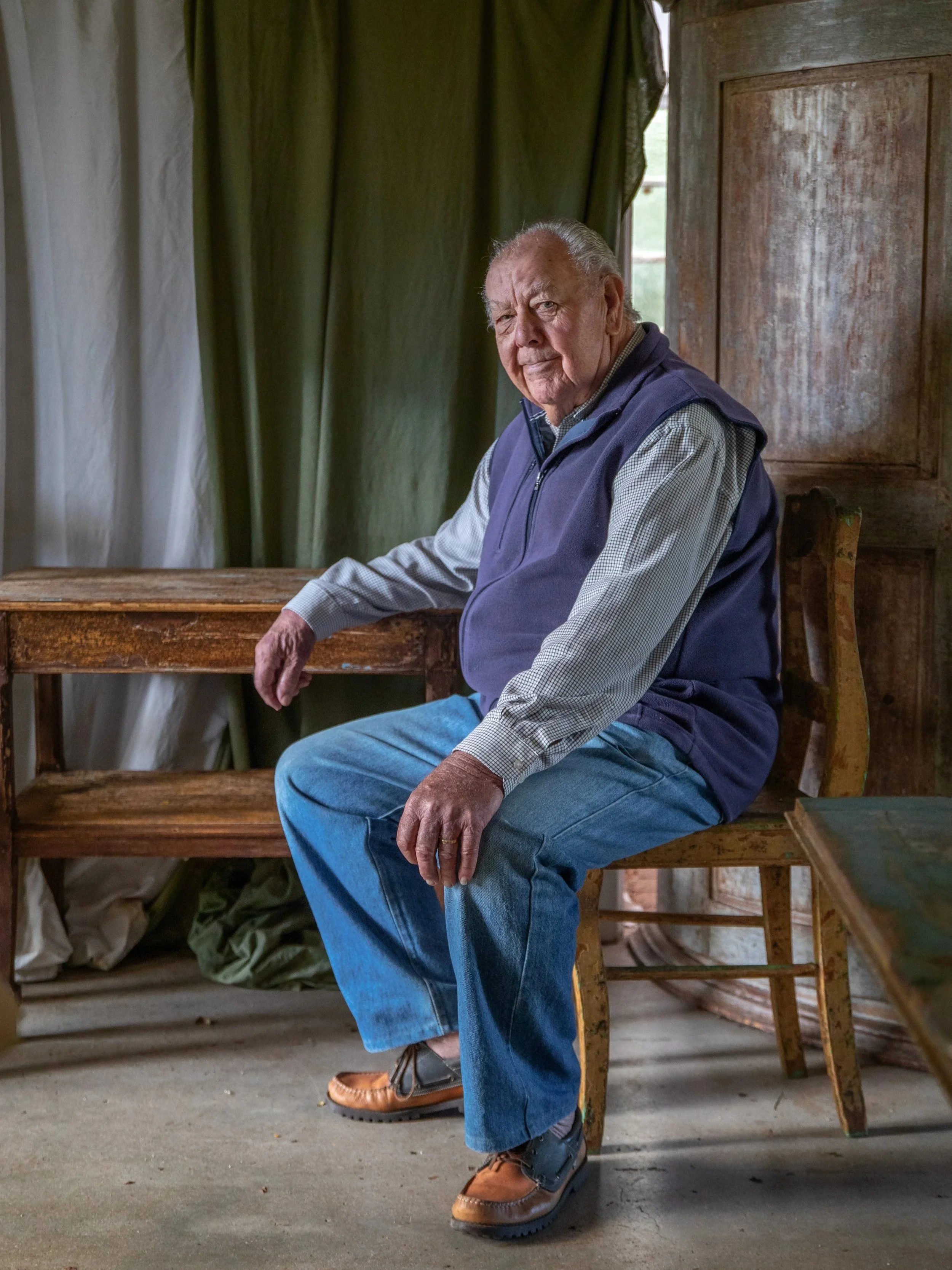 An elderly man with gray hair sitting on a wooden bench, wearing a blue vest, long-sleeved shirt, jeans, and brown shoes, in a rustic room with dirt floor, wooden walls, green curtains, and natural light coming through a window.