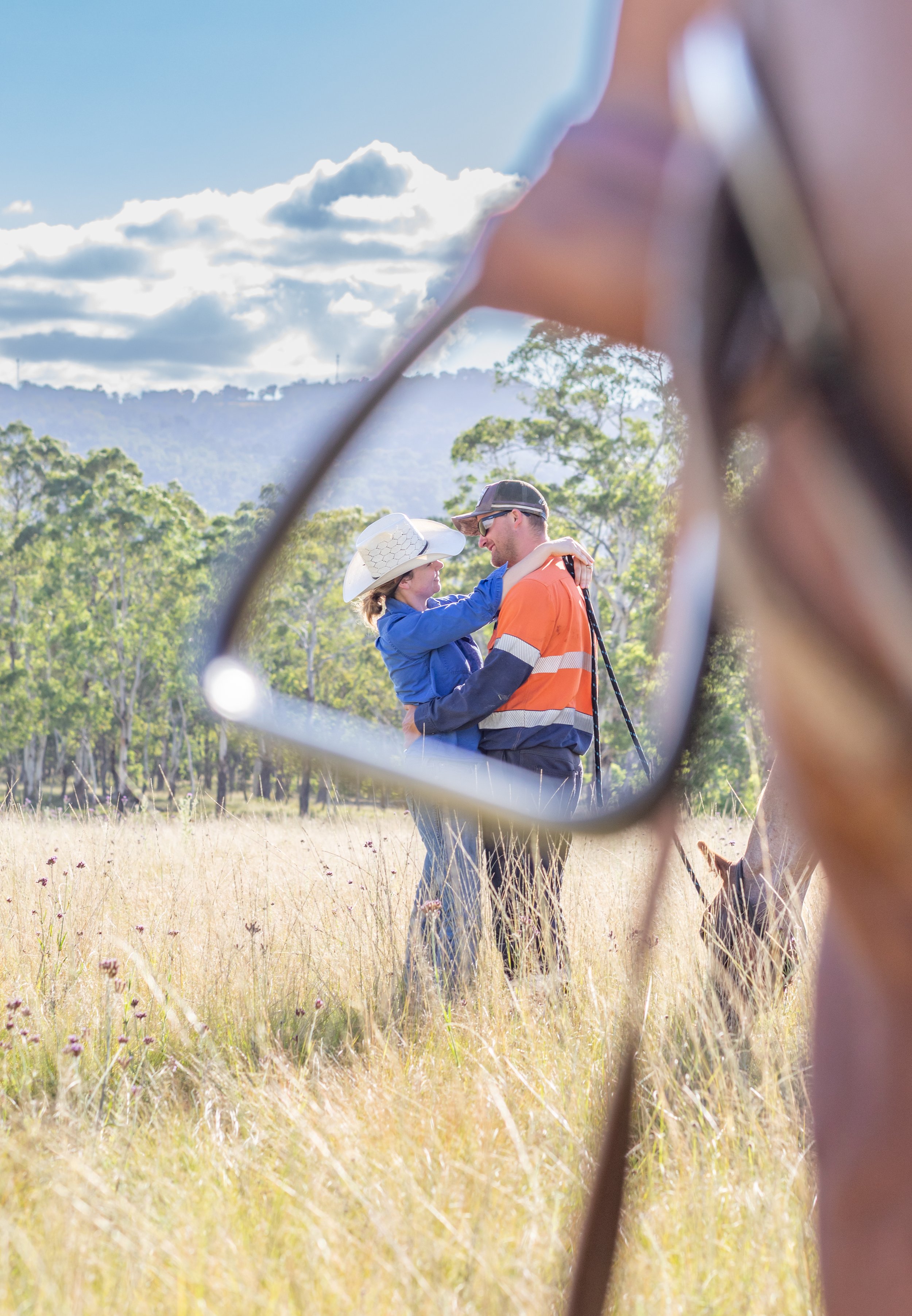 A couple dancing in a field is seen through the reflection of glasses, with lush trees and a cloudy sky in the background.