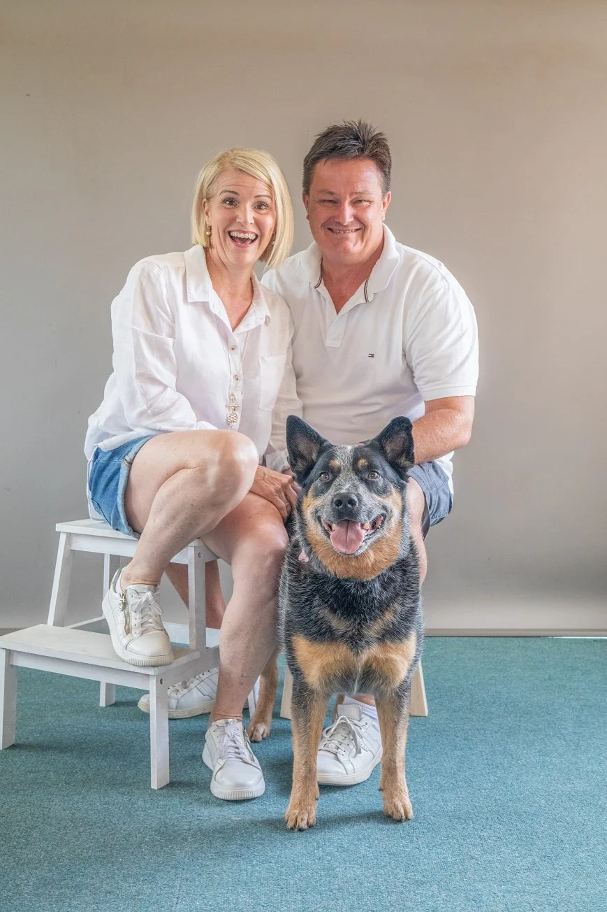 A smiling blonde woman and a man sitting on a white chair with a dog in front of them, all dressed casually in white shirts and shorts, in a room with a plain beige wall and teal carpet.