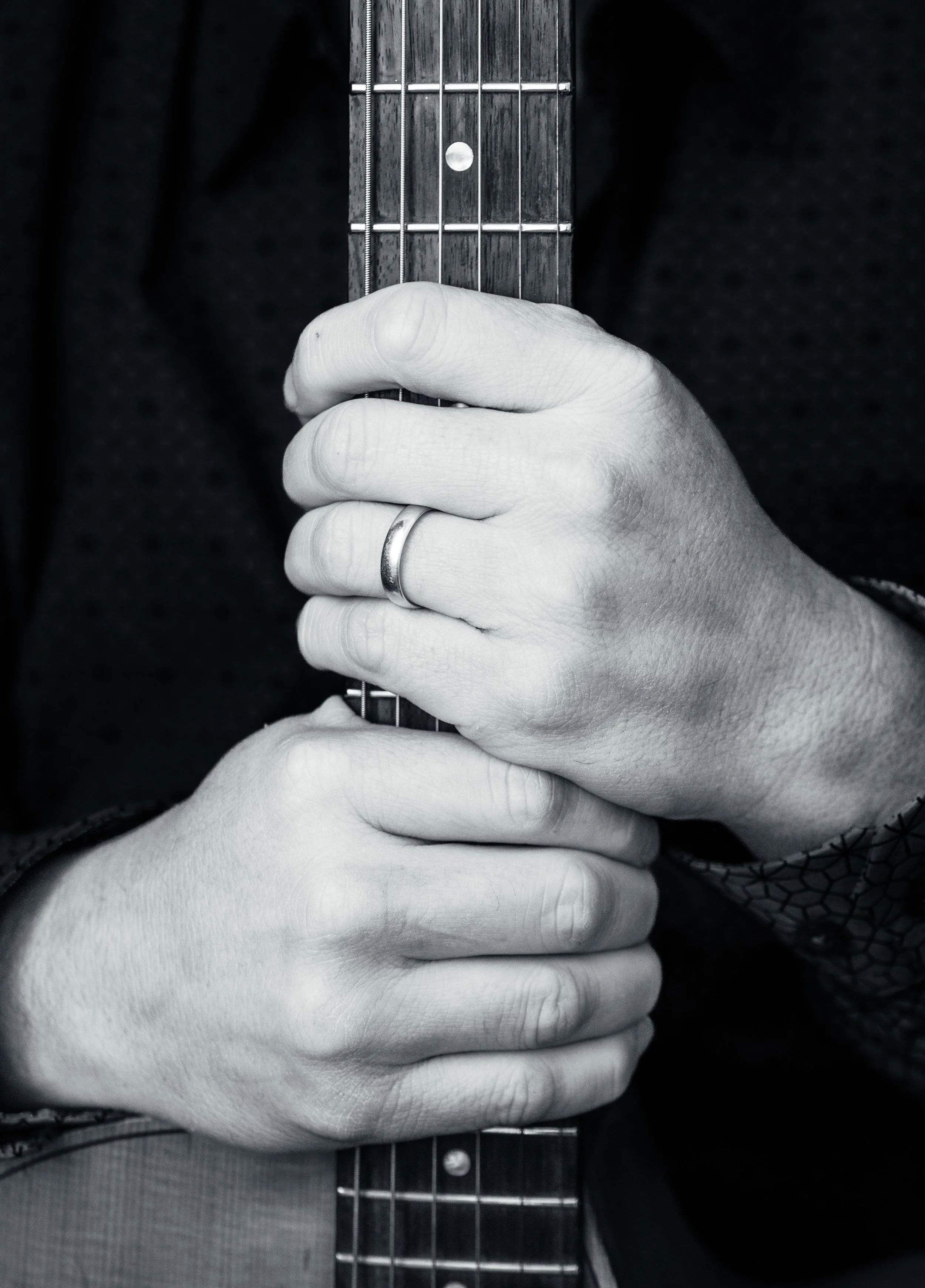 Close-up of a person's hands gripping the neck of an acoustic guitar, with a wedding band on the left ring finger, in black and white.