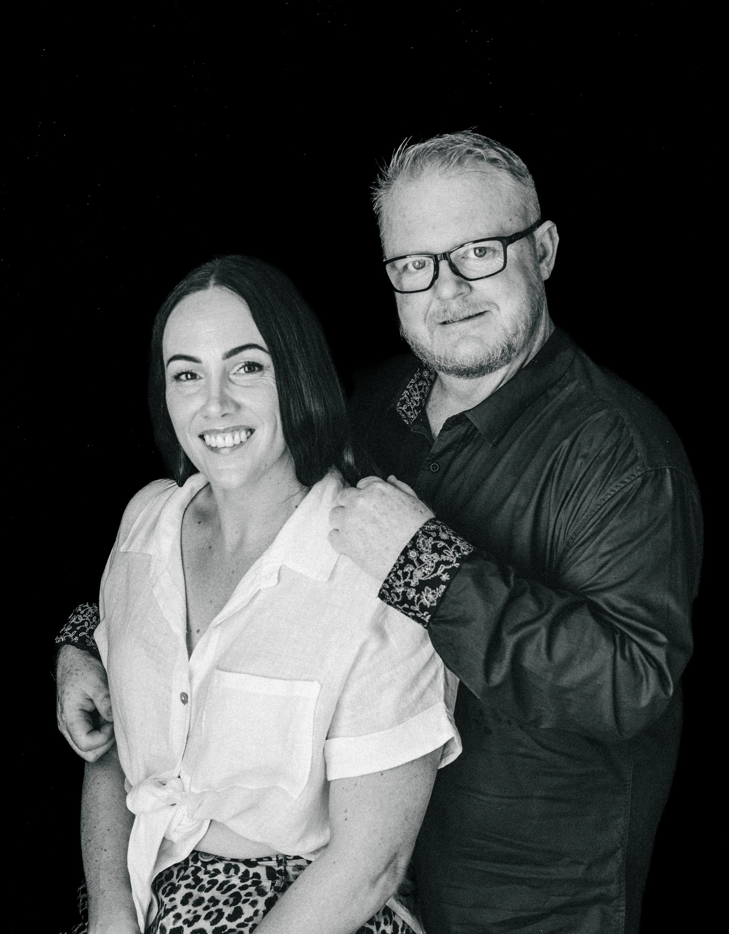A man and woman pose together against a black background. The woman has straight dark hair, a white shirt with rolled-up sleeves, and leopard-print pants. The man has light hair, glasses, and a beard, and is wearing a dark shirt with embroidered cuff