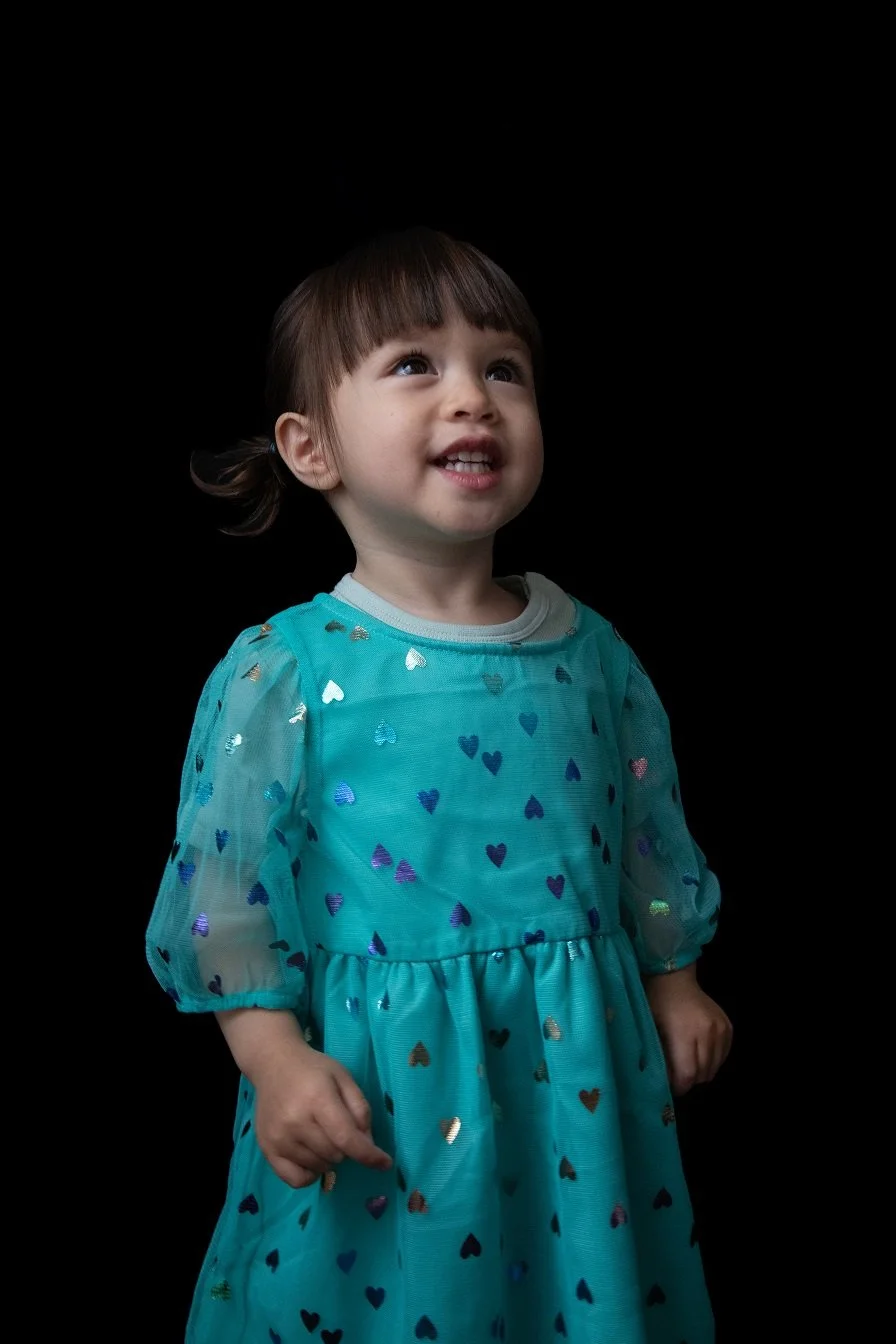 A young girl with brown hair and bangs, wearing a turquoise dress with multicolored heart patterns, looking upward with a happy expression against a black background.