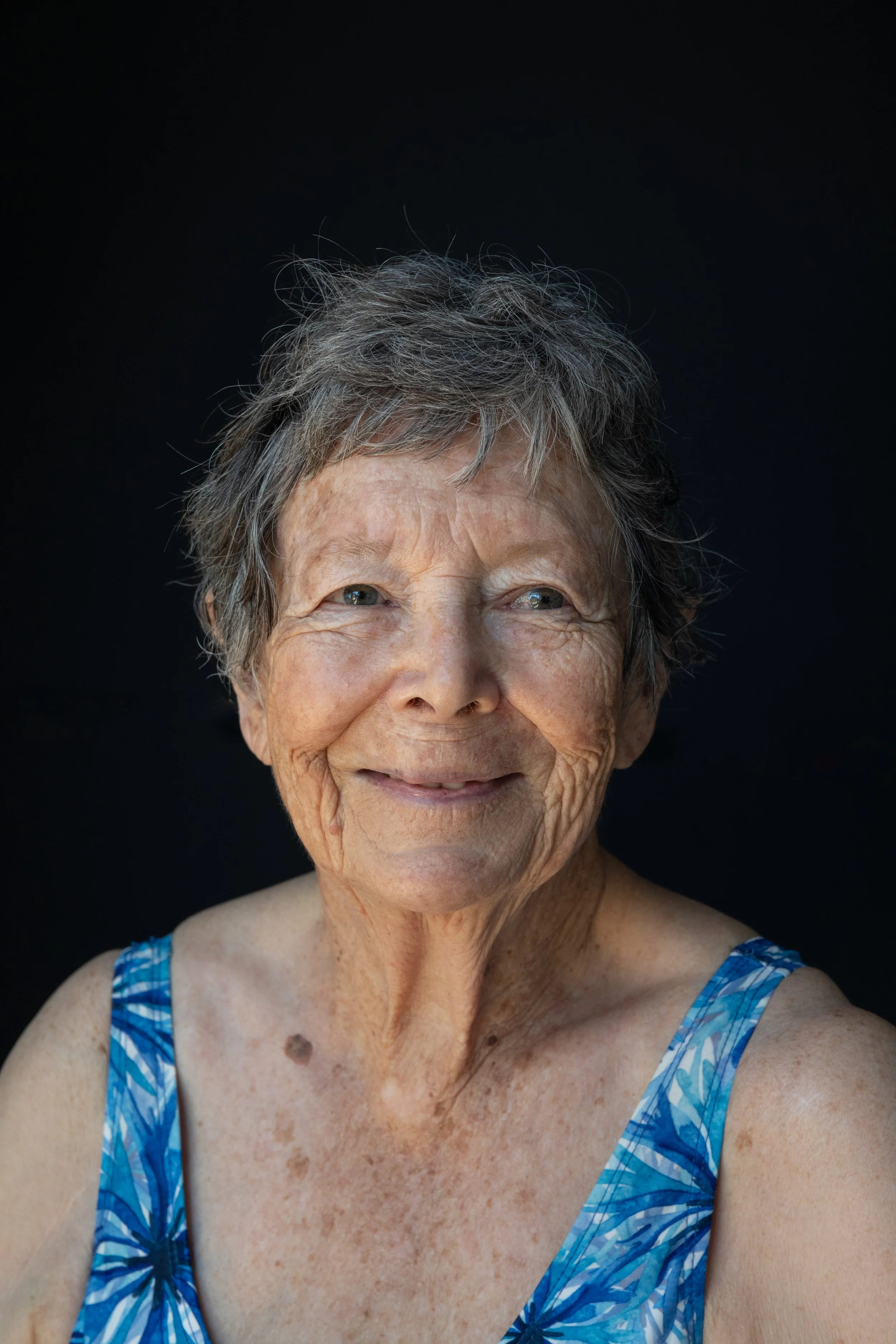 Close-up portrait of an elderly woman with short gray hair, wearing a blue floral sleeveless top, smiling gently against a black background.